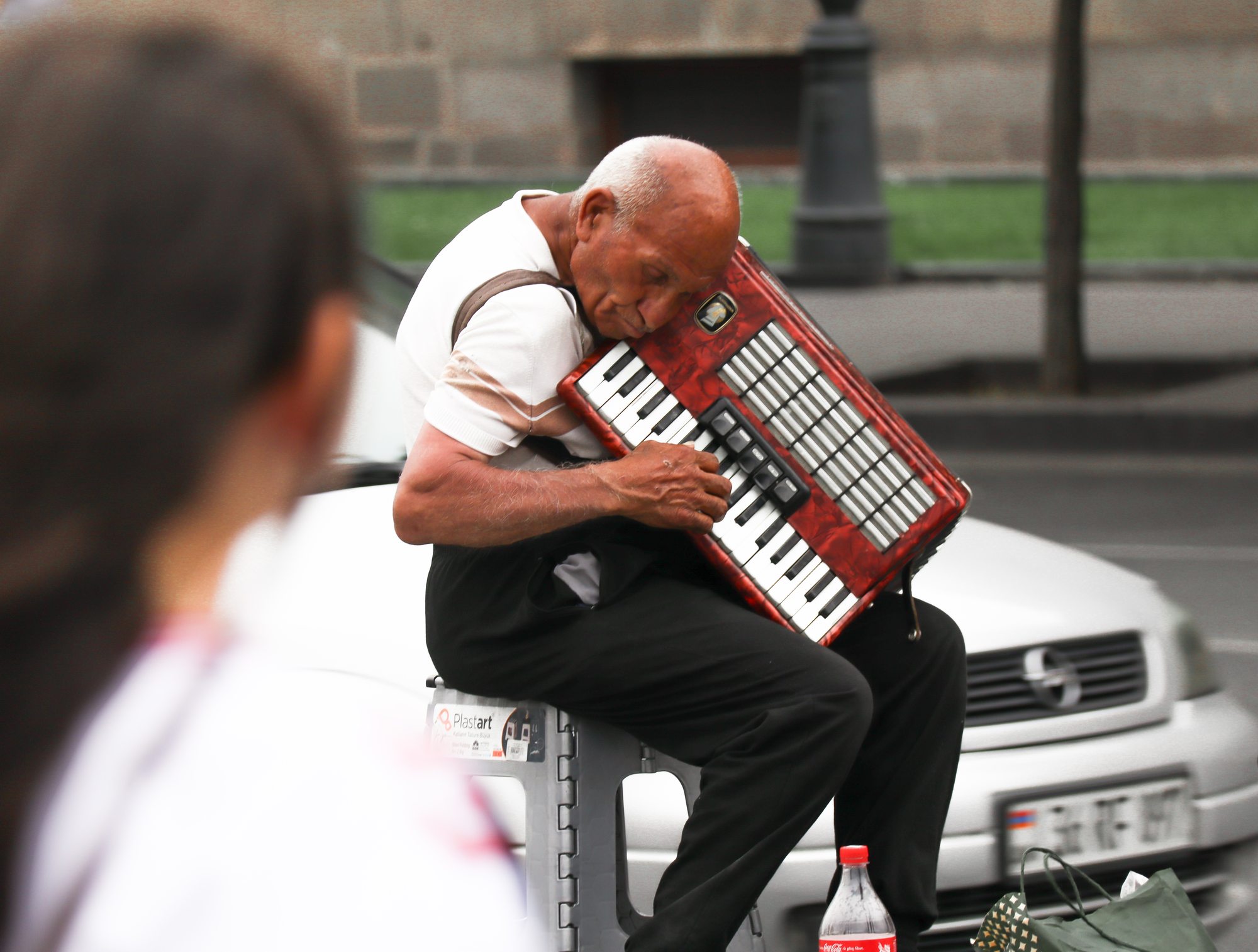An elderly man playing accordion on a Yerevan street corner — the warm tuff stone of the buildings glowing behind him in the afternoon light