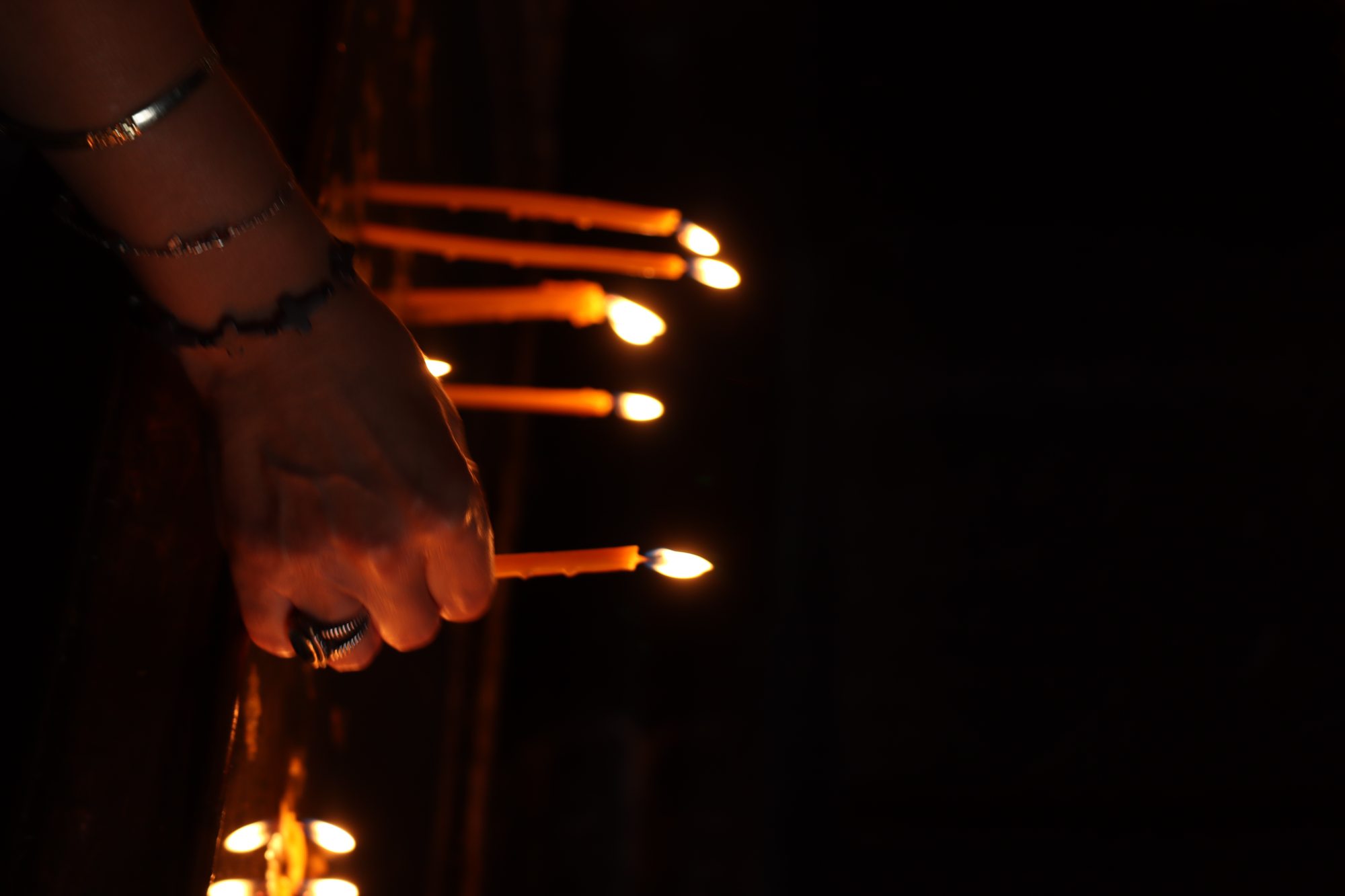 A hand lighting candles in the deep darkness of an Armenian church — flame reflected in the ring on the finger, the dark stone of the church behind