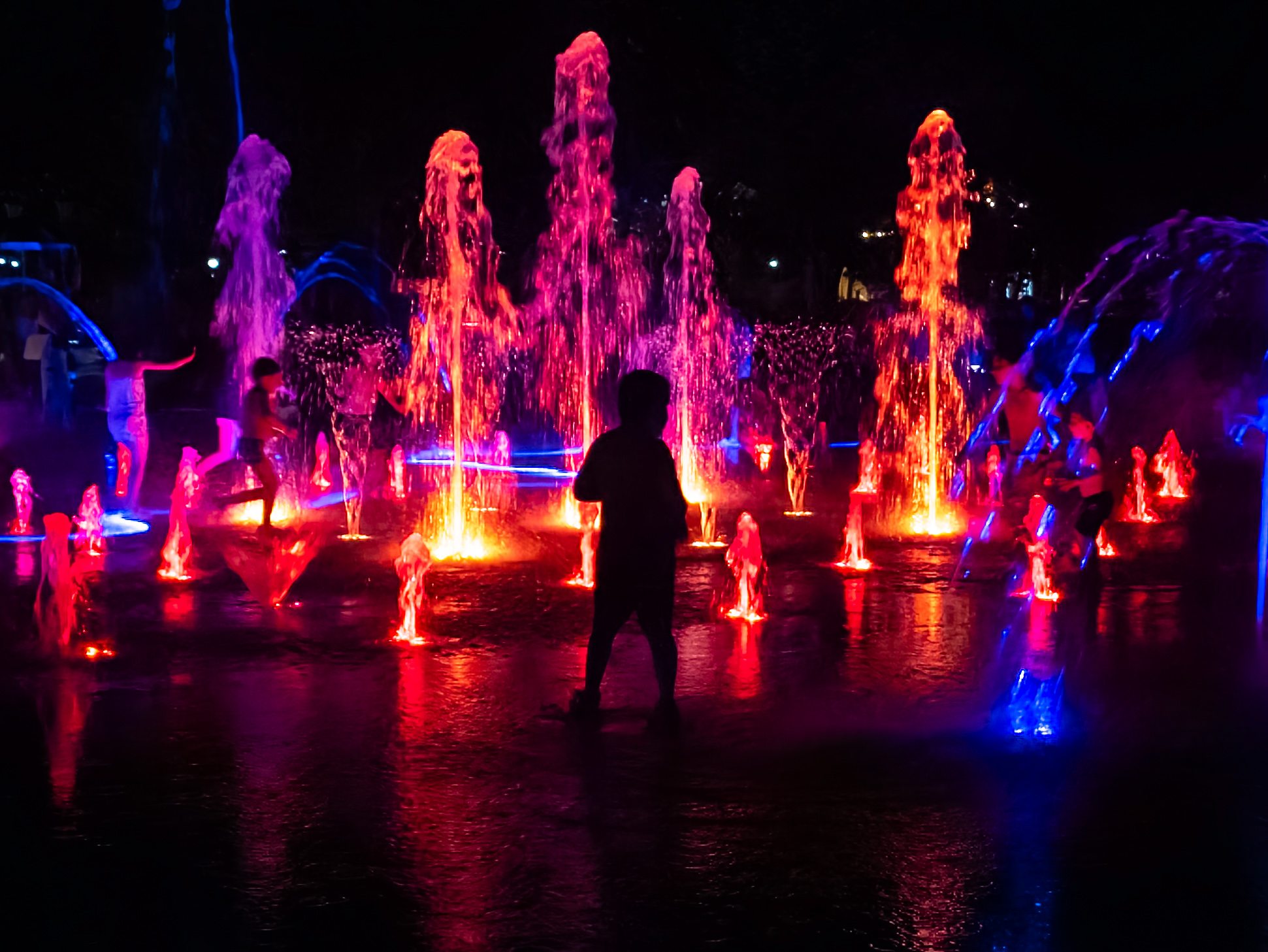 Republic Square in Yerevan at night during Vardavar — children playing in the illuminated fountains, silhouettes against the red and blue light, the square alive