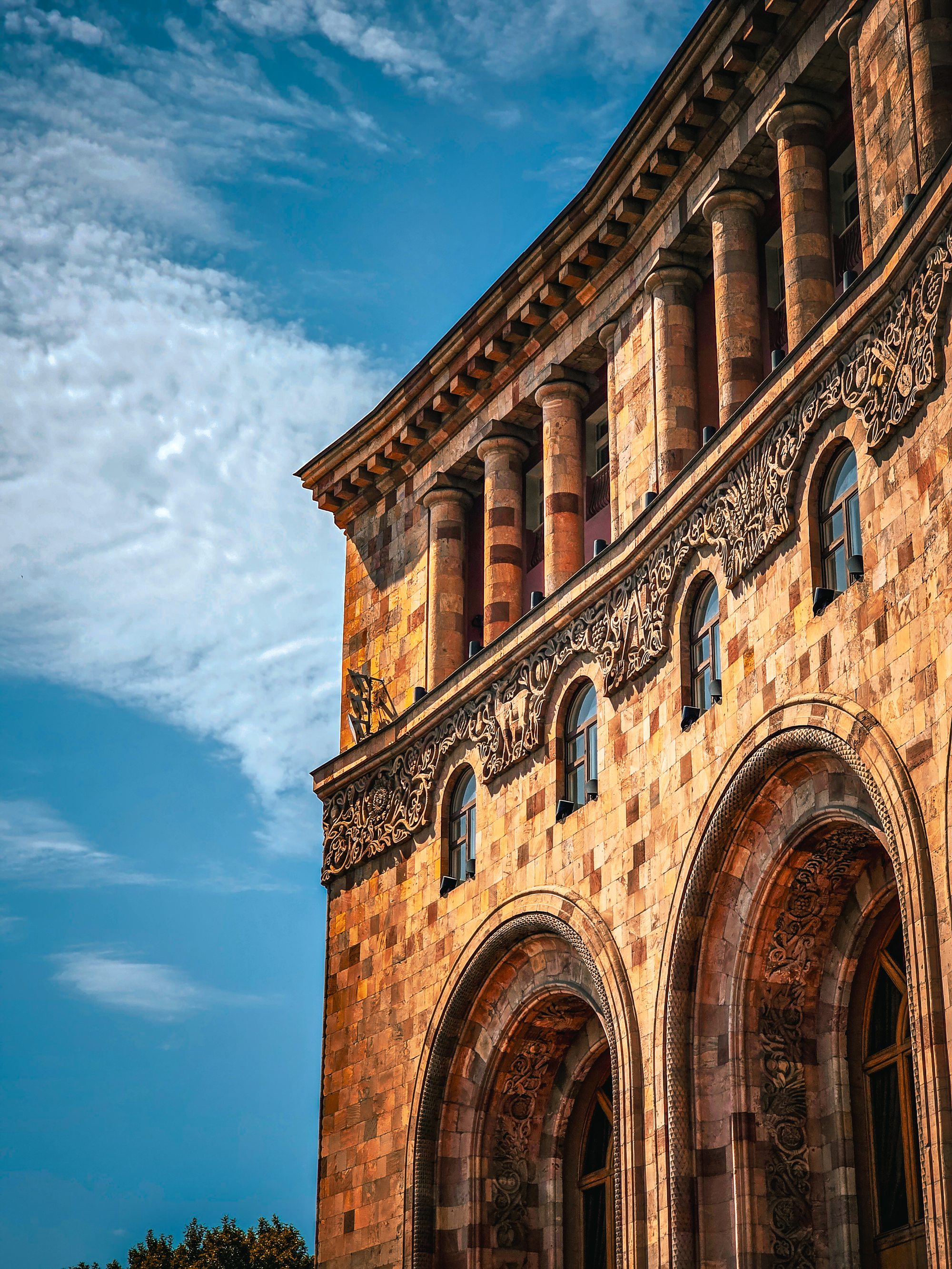 The intricate carved facade of a Yerevan tuff stone building at golden hour — centuries of Armenian architecture glowing warm pink against a blue sky