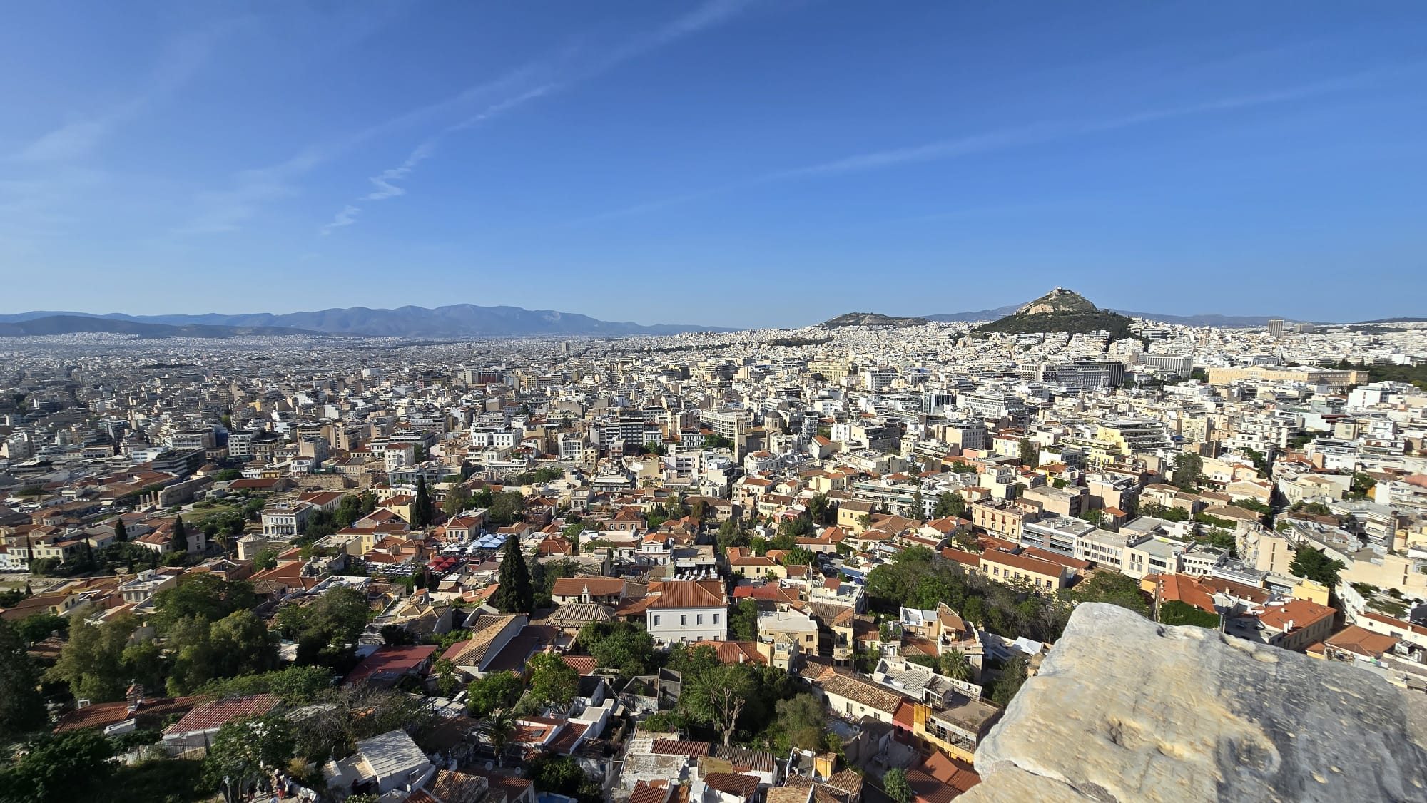 A panoramic view across the Athens cityscape — dense white and terracotta buildings stretching to the distant hills