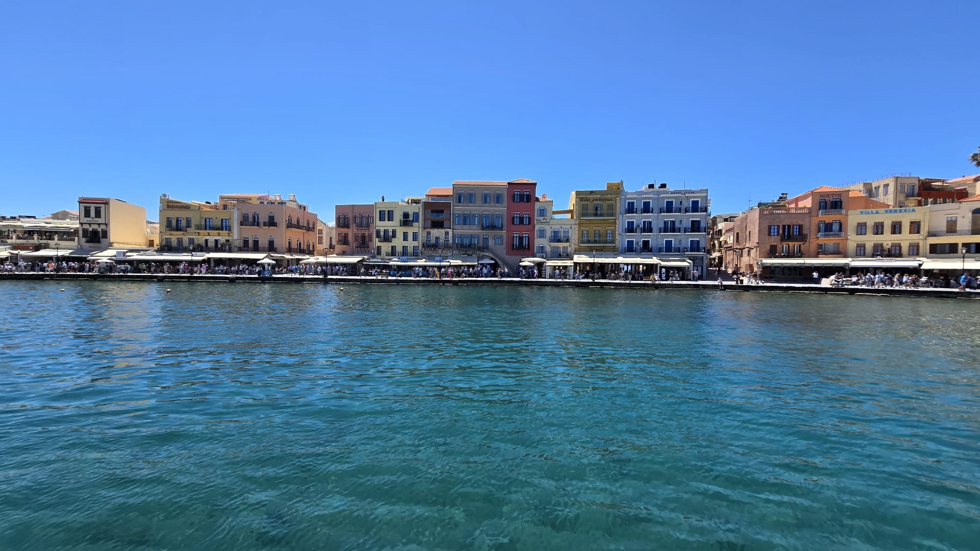 The coastline of Crete from a tour boat — clear turquoise water against rocky shore under a hot blue sky