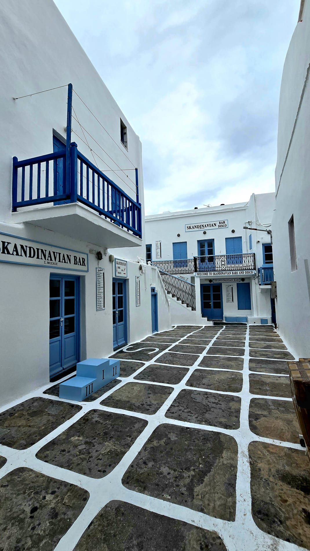 A winding whitewashed alley in Mykonos town, blue-painted steps and church dome visible at the end