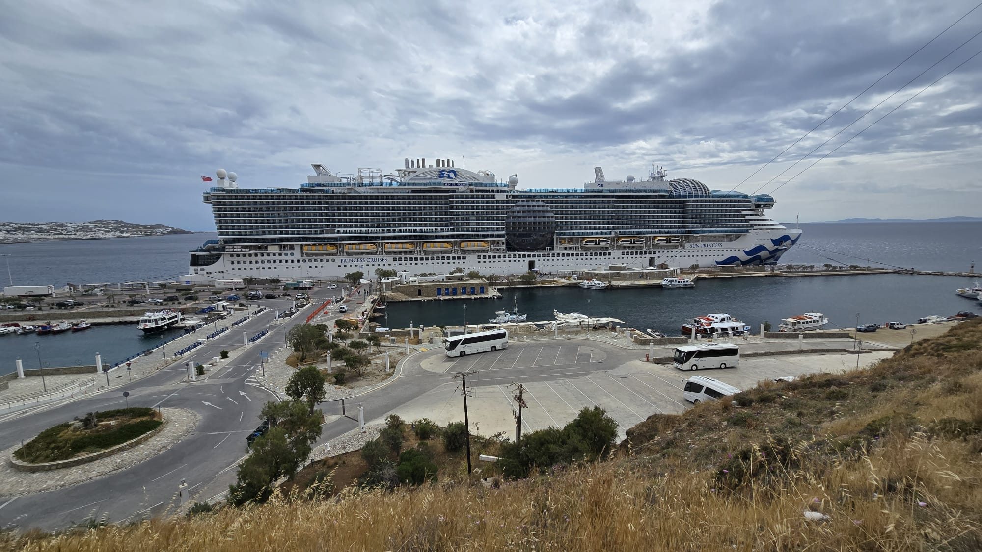 A large cruise ship anchored in the blue water off the new port of Mykonos, seen from the coastal road