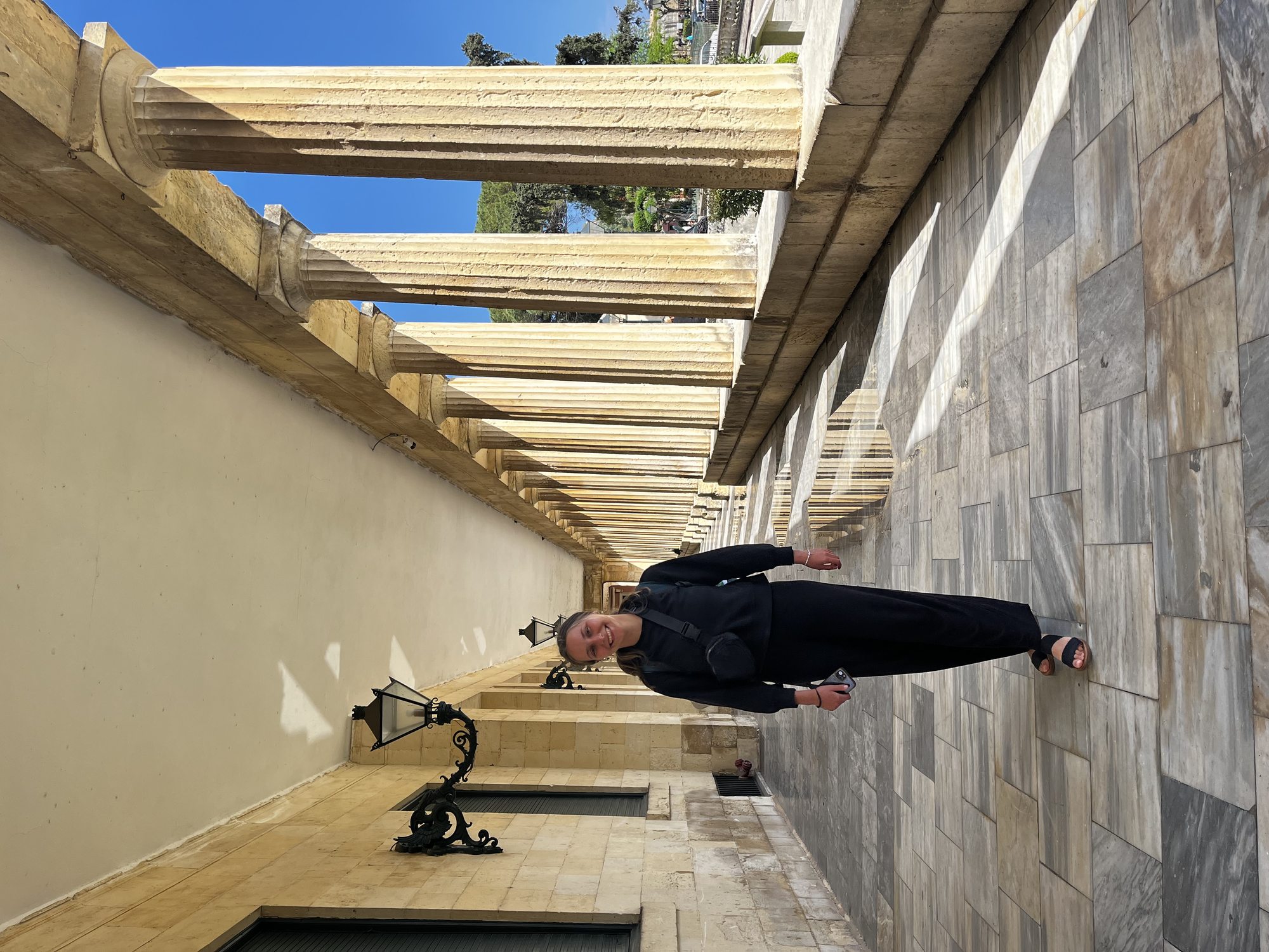Yaron in dark clothes standing under a colonnade in Corfu Old Town — large square stone columns and beam ceiling overhead casting deep shadows on a tiled walkway, an iron lantern visible above, marble wall on the right