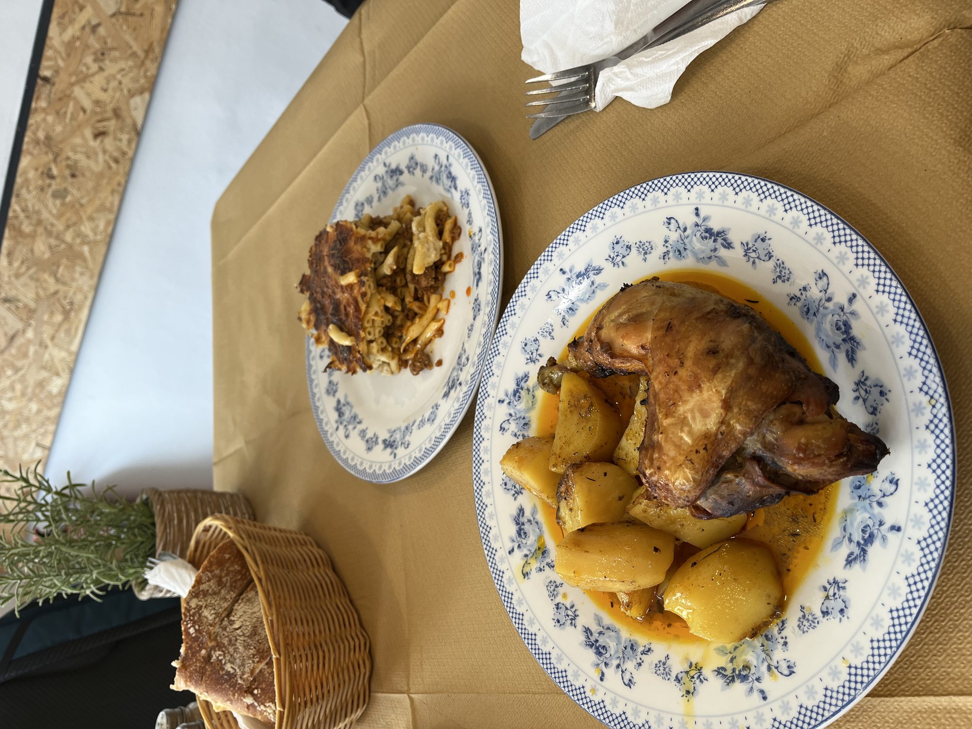 A homemade Greek-Italian meal on a wooden table — a large blue-and-white plate of roasted chicken with golden potatoes in a pan sauce, a side plate of cabbage salad, a bread basket on the side, brown paper tablecloth