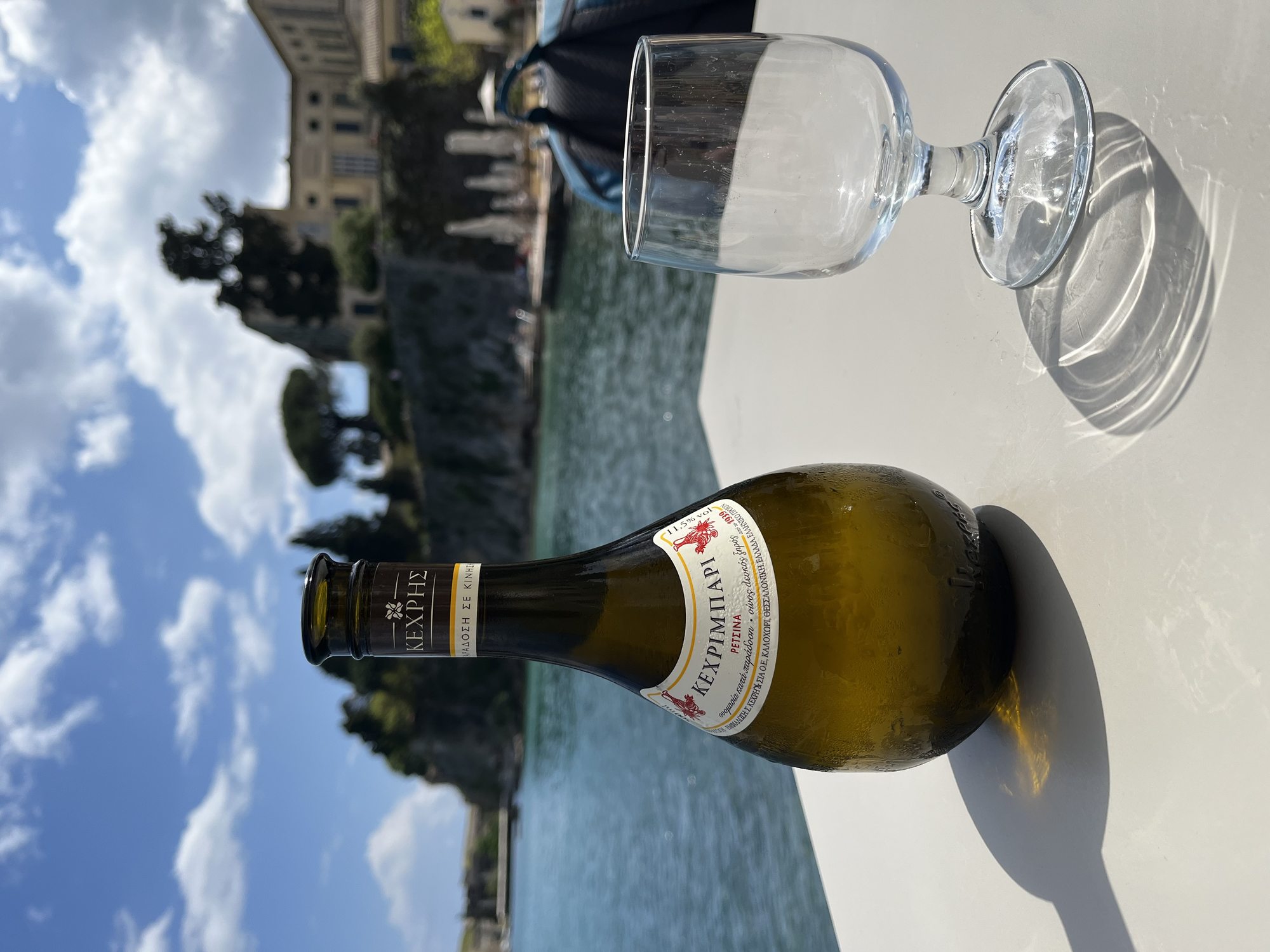 A bottle of Kechribari Corfiot white wine and an empty wine glass on a white café table, with the Old Fortress of Corfu Town visible across the water in the background under a blue sky with white clouds