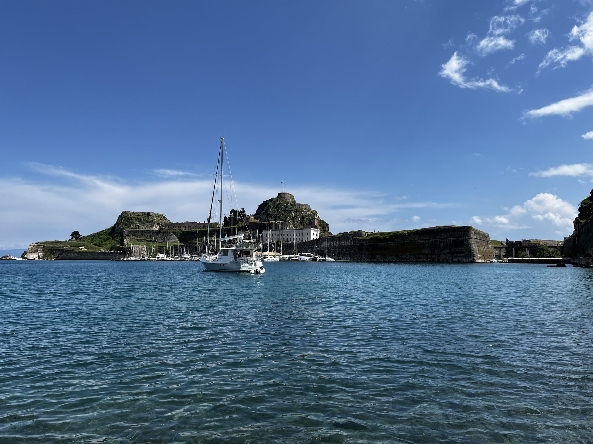 A wide view of the Old Fortress of Corfu from across the harbour — long Venetian stone walls running along a rocky promontory, two hills behind them, a sailboat moored in the foreground on still blue water under a wide blue sky
