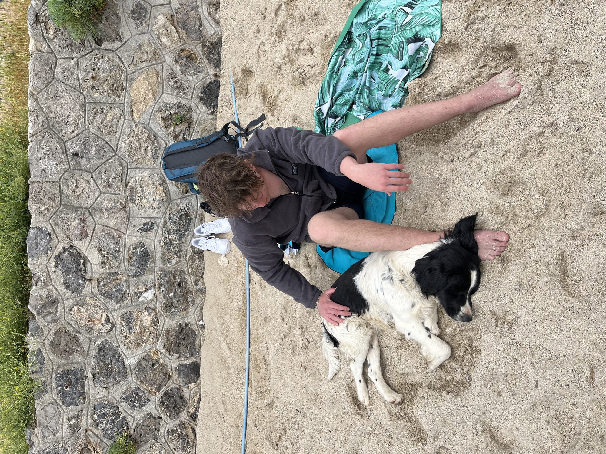 Yaron sitting on a beach towel on the sand with a black and white dog lying calmly beside him, a backpack and water bottle next to them, stone slabs framing the edge of the sand