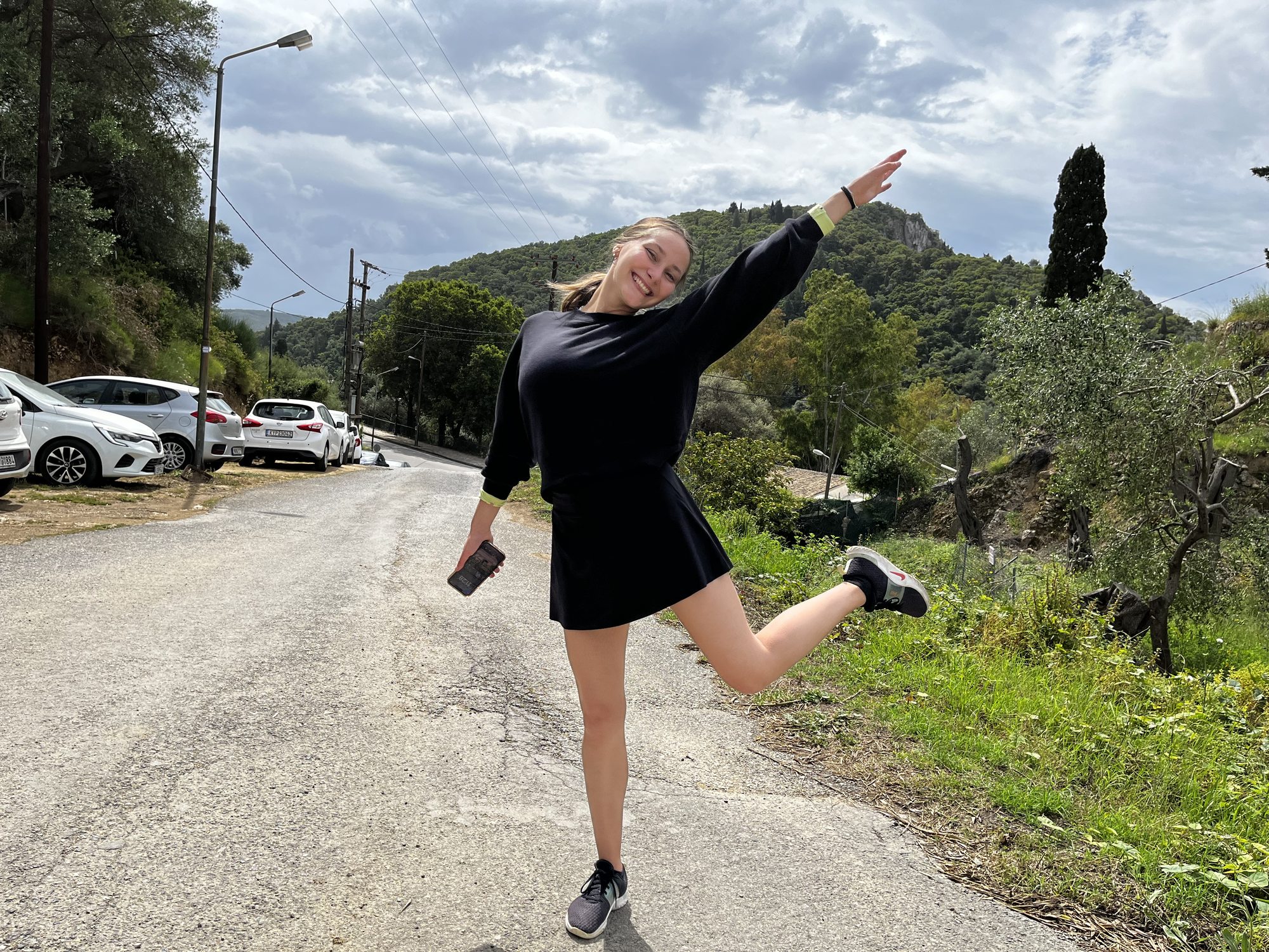 Late afternoon at the entrance to Paleokastritsa — a small road climbing past a stone wall and a hillside of green olive and cypress trees, with a wedge of distant turquoise sea visible past the hill, a young woman in a black sweatshirt and skirt mid-skip on the road with arms up