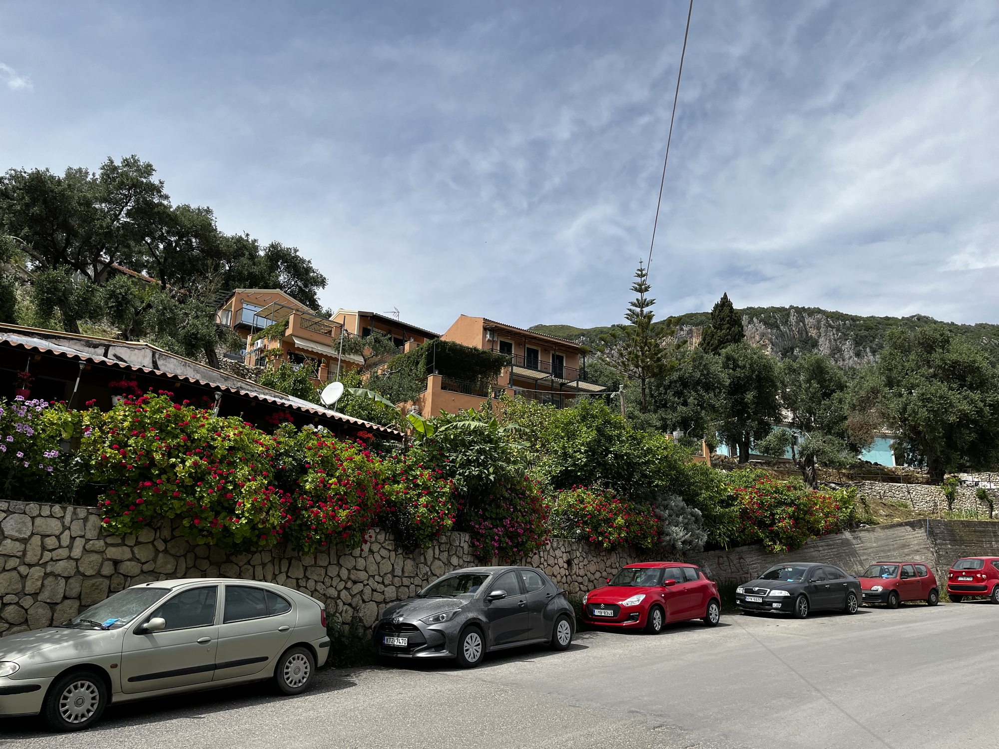 A village street above Paleokastritsa with parked cars, salmon-pink and ochre houses, terracotta roofs, and walls of cascading red bougainvillea, mountains visible in the distance