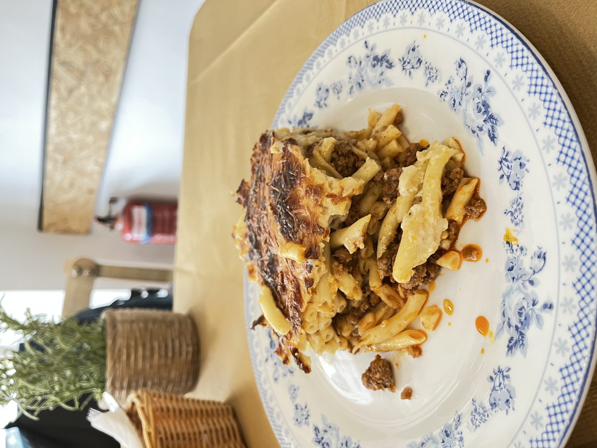 A thick wedge of homemade Corfiot pastitsio on a blue-and-white plate — long pasta tubes layered with cinnamon-tomato meat sauce and a deeply browned béchamel top, a sprig of rosemary on the table beside it