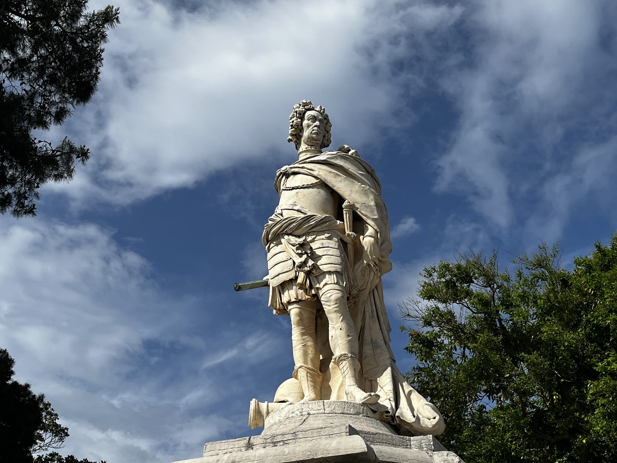 A white marble statue of a man in Roman-style military armour with a sword at his side standing on a stone plinth, framed by deep green trees and a partly cloudy blue sky