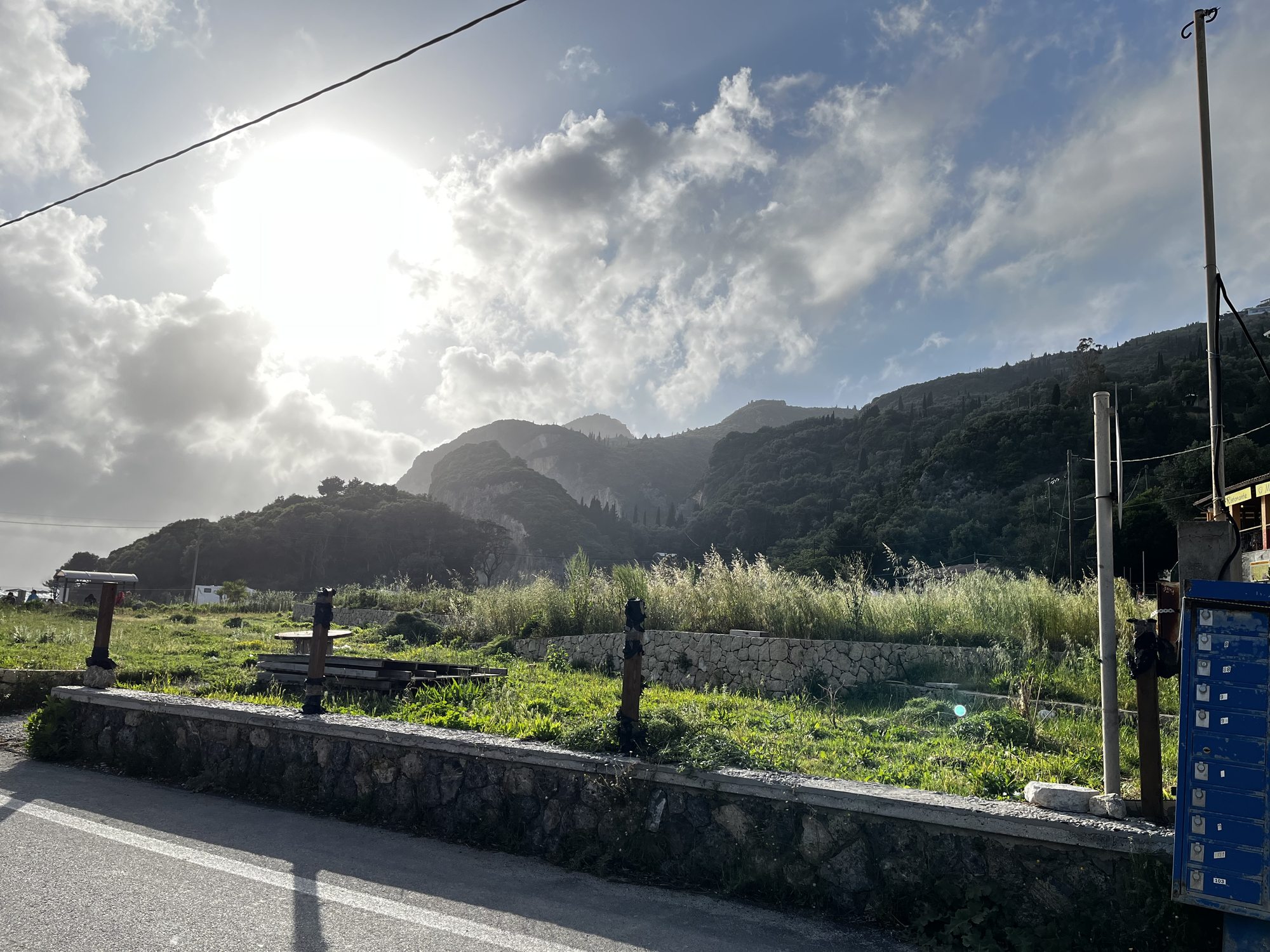 Evening light at Paleokastritsa with the sun behind a ridge of green hills, scattered houses on the slope, low scrub and grass in the foreground, a stone wall along the road, light cloud cover above