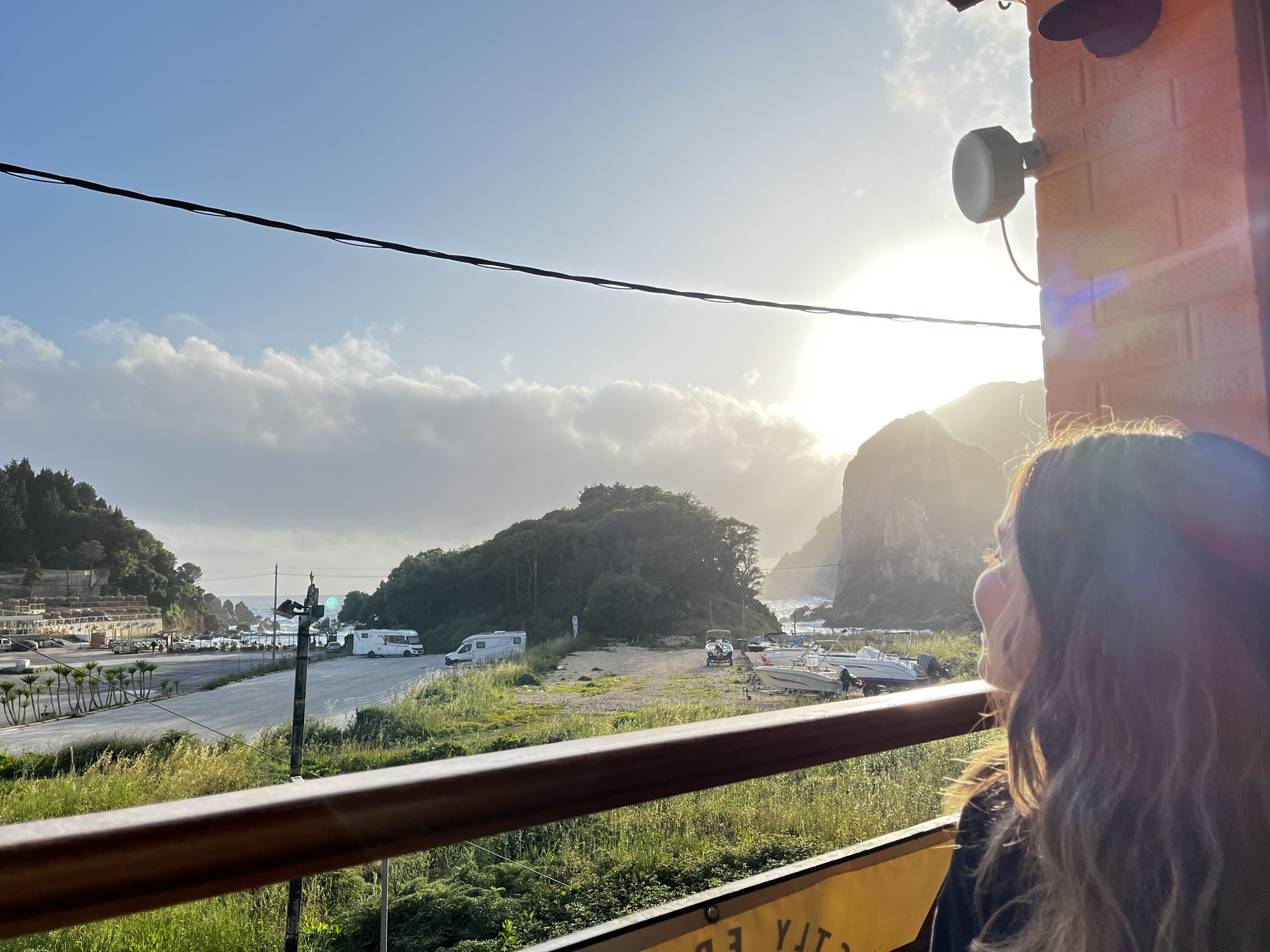 A view from a wooden terrace at golden hour overlooking the Paleokastritsa bay — a green headland and cliffs catching the last sun, a small marina with boats and a few campervans in the parking lot below, the silhouette of a woman with long hair in the foreground looking out