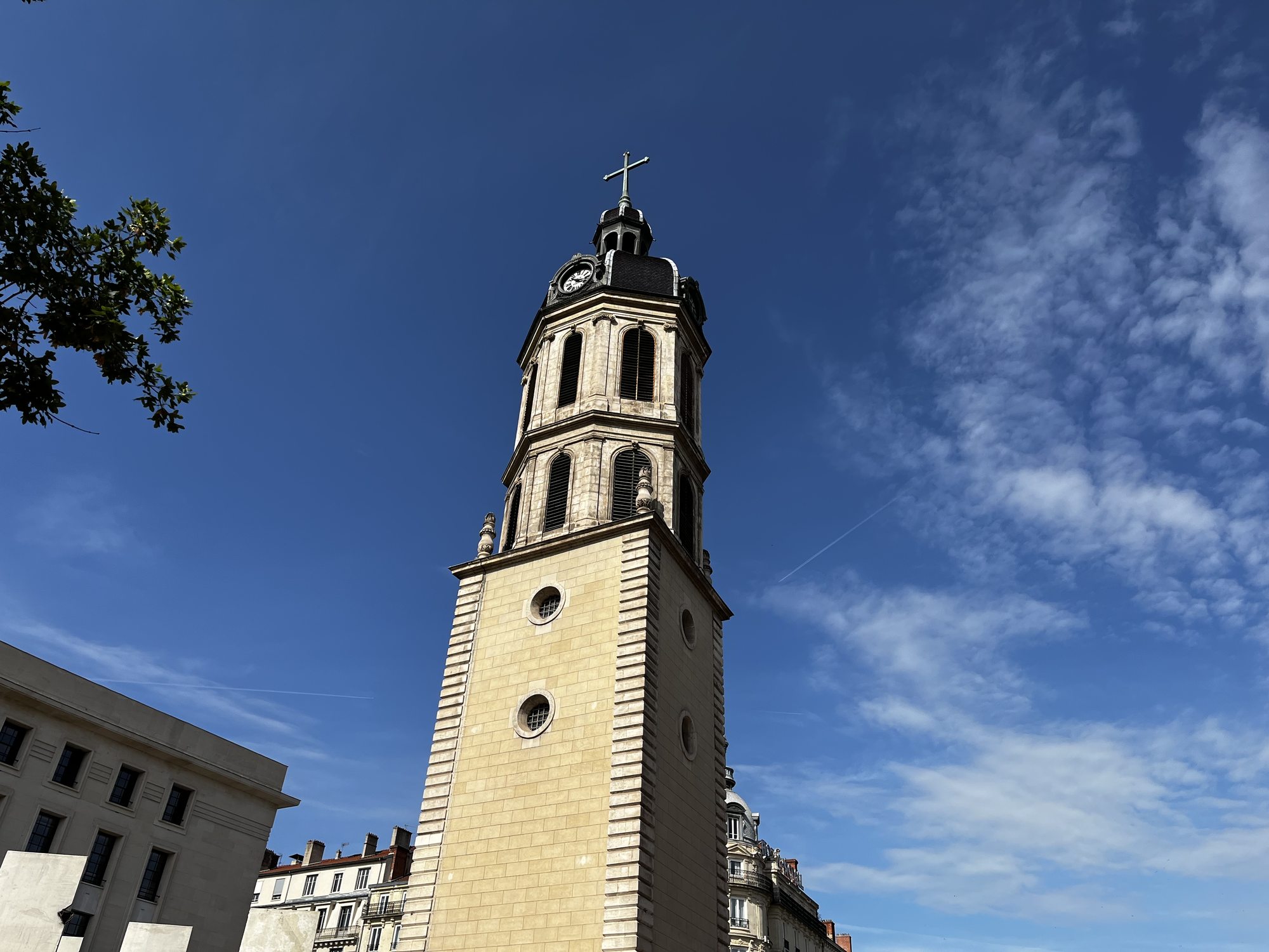 A 17th-century bell tower in central Lyon photographed from below against a deep blue sky with cirrus clouds — the clock face and cross visible at the top