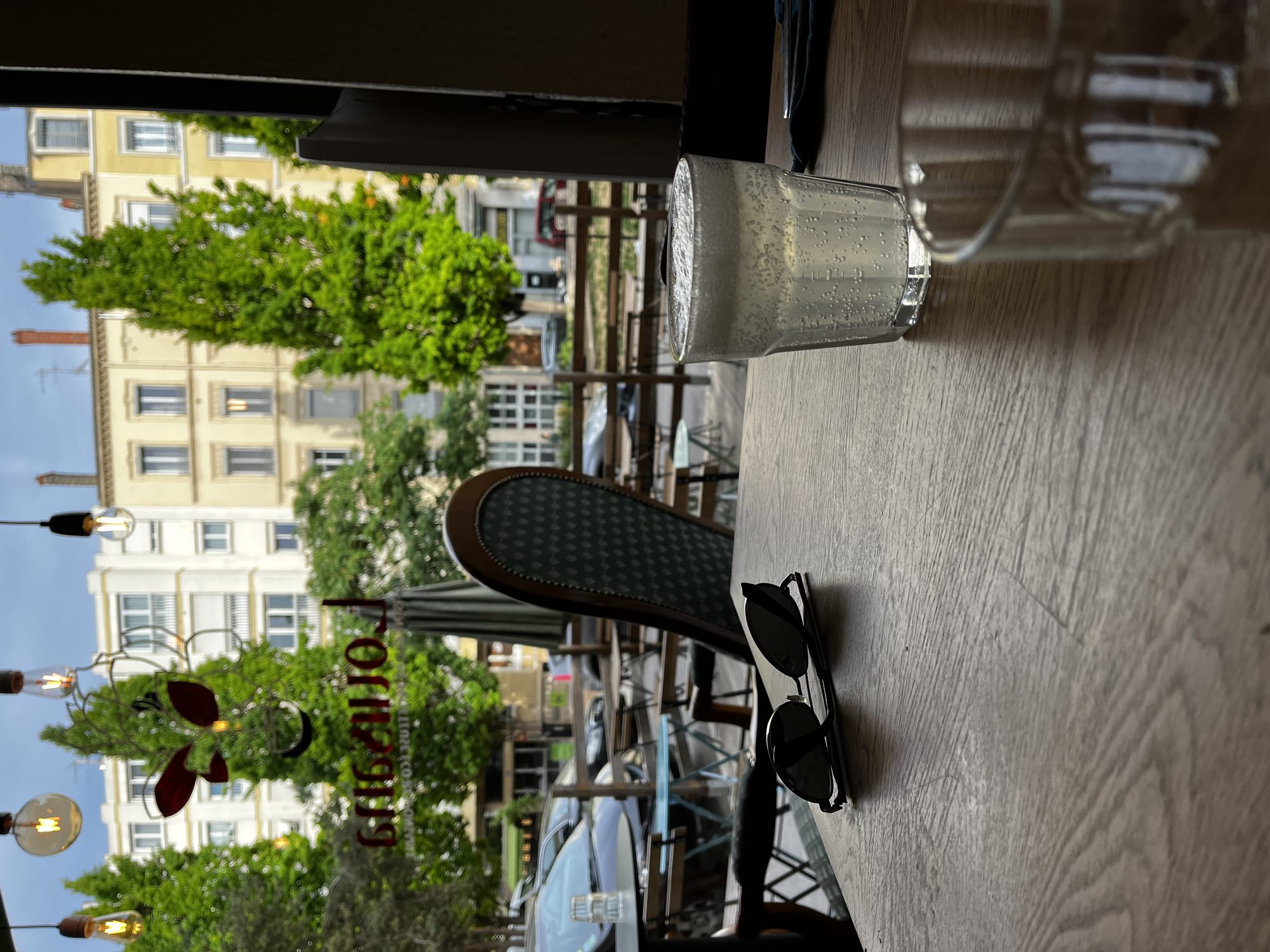 A café terrace on the Presqu'île in Lyon at morning — sunglasses on a wooden table, an aperitif glass, the square and a Lyon façade beyond