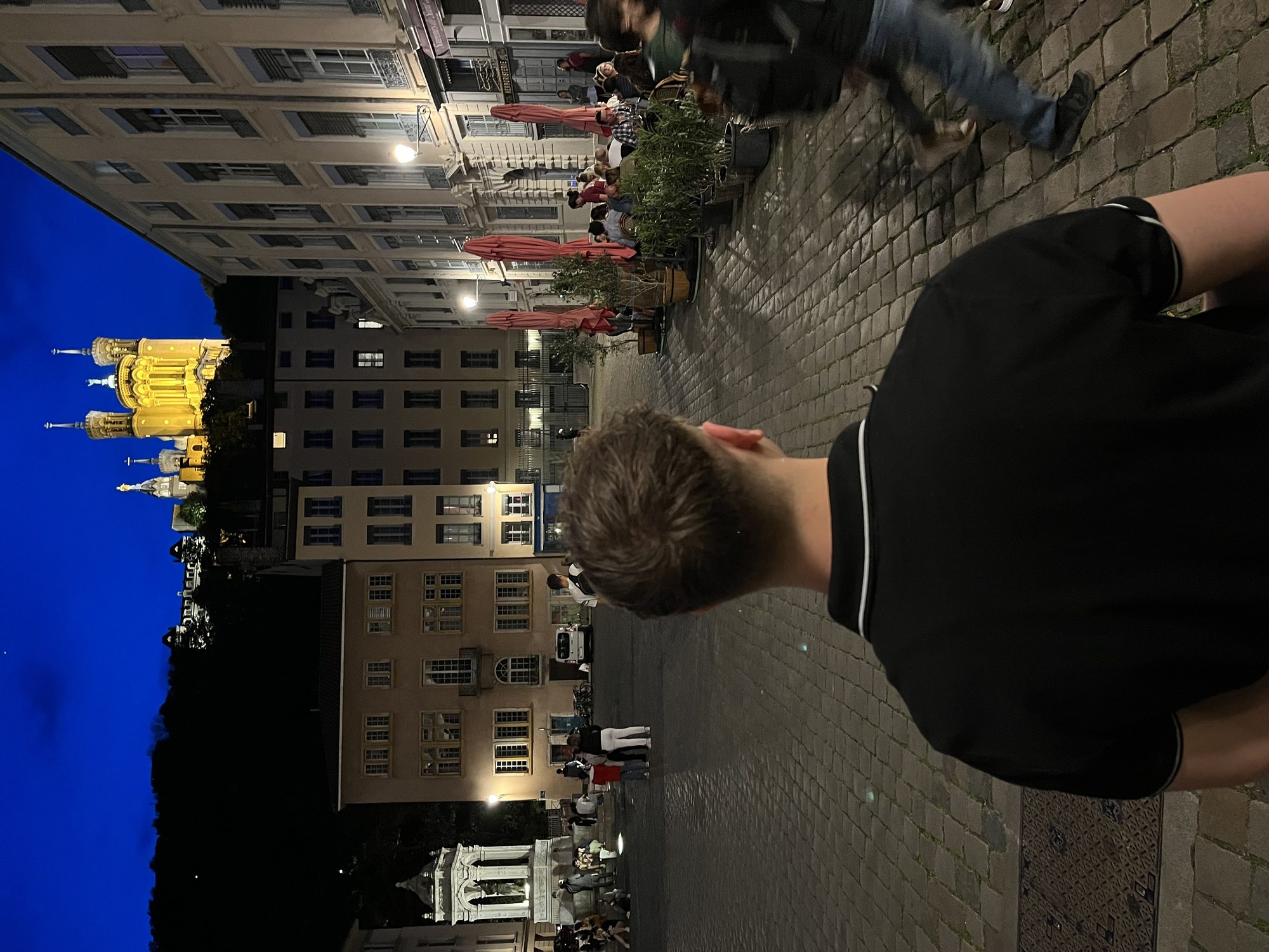 Yaron at Place des Terreaux in Lyon at night — the illuminated golden Bartholdi fountain visible behind him, café terraces lit in soft warm light
