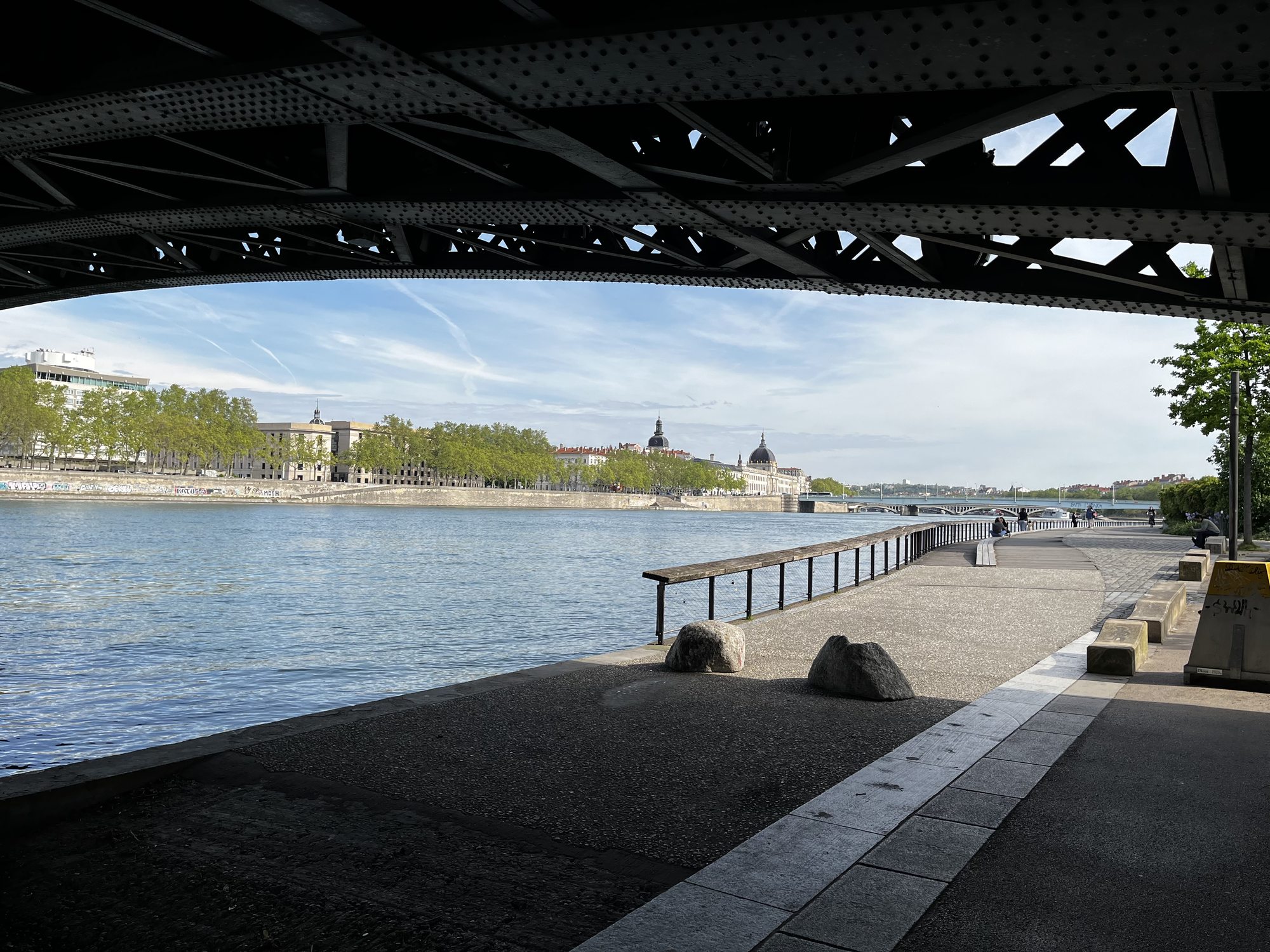 A view from under a steel railway bridge along the Saône riverbank in Lyon — looking back toward the Hôtel-Dieu dome and the historic waterfront