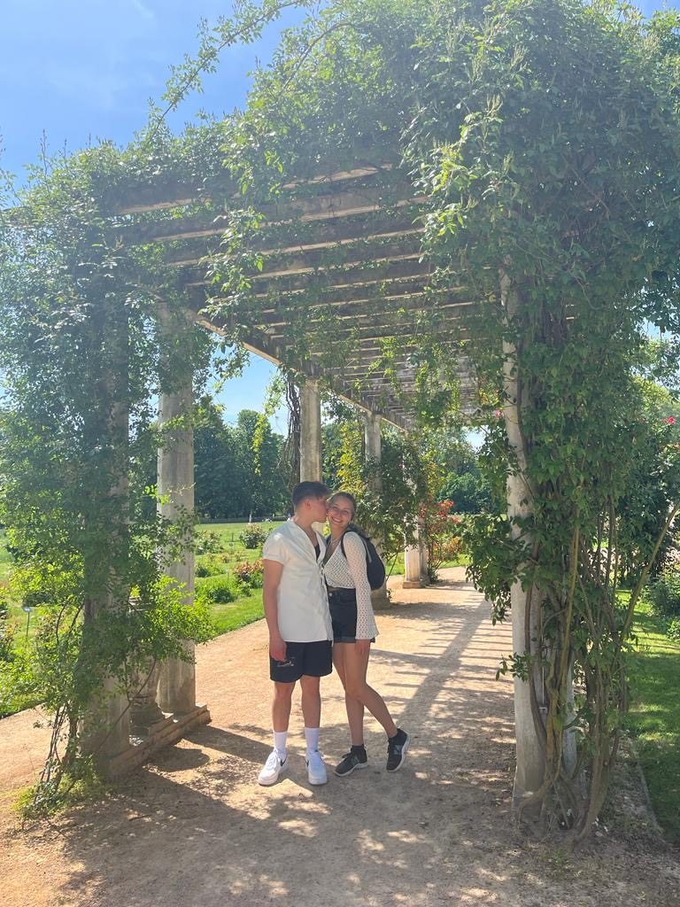 A couple posing under a wisteria-covered wooden pergola at Parc de la Tête d'Or in Lyon — long shafts of sunlight, the rose garden behind them