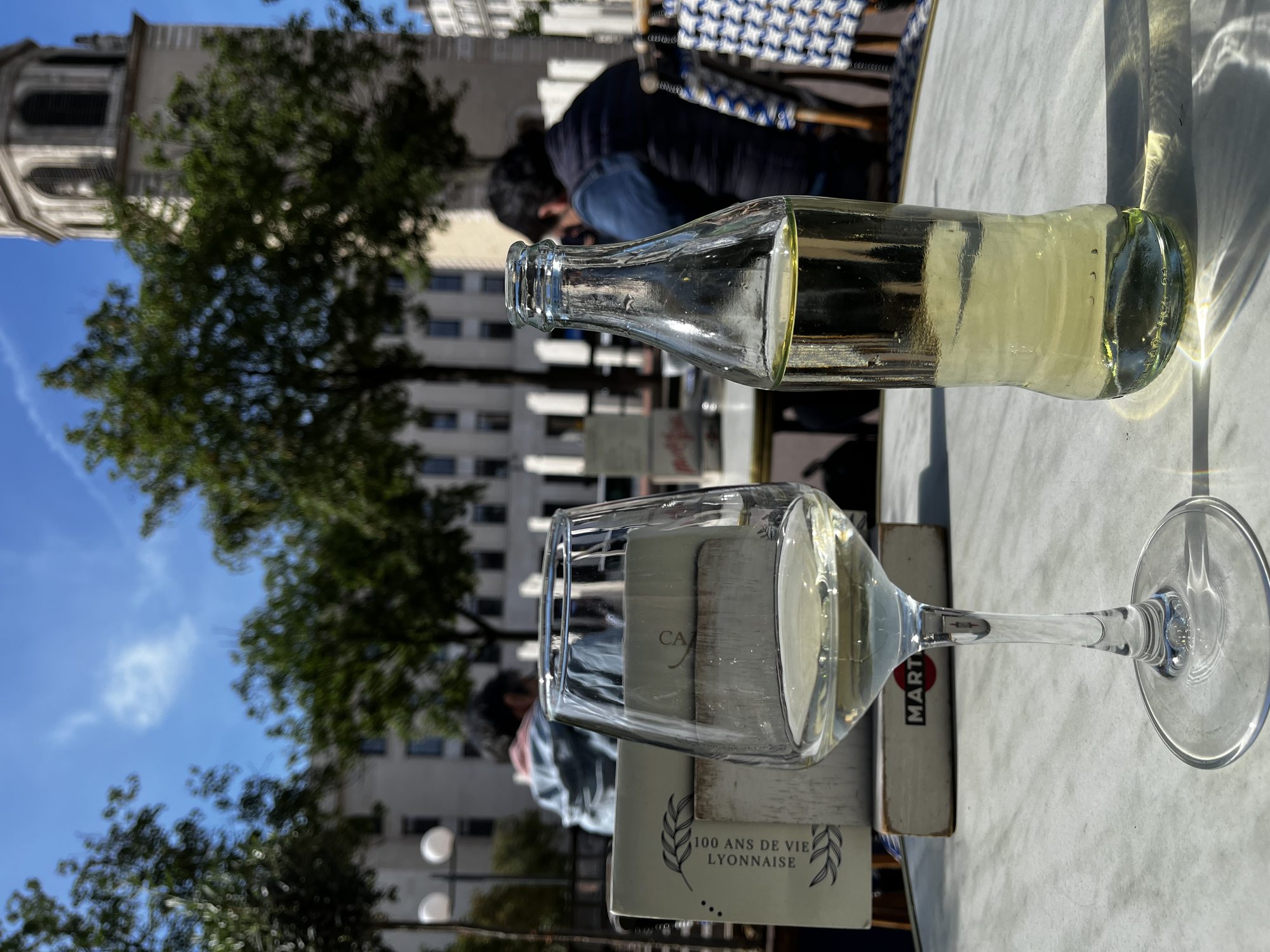 A carafe of white wine and a champagne glass on a marble café table in Lyon — a napkin holder marked 100 ANS DE VIE LYONNAISE