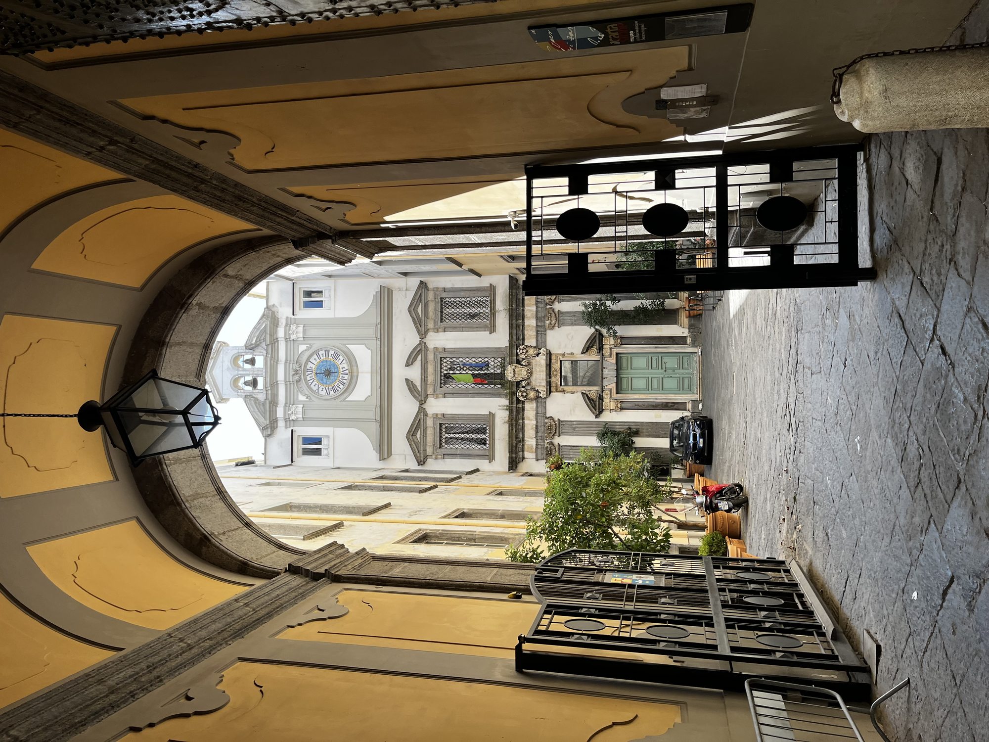 An ornate baroque archway and inner courtyard in the centre of Naples — frescoed ceiling, wrought-iron details and a glimpse of the city beyond