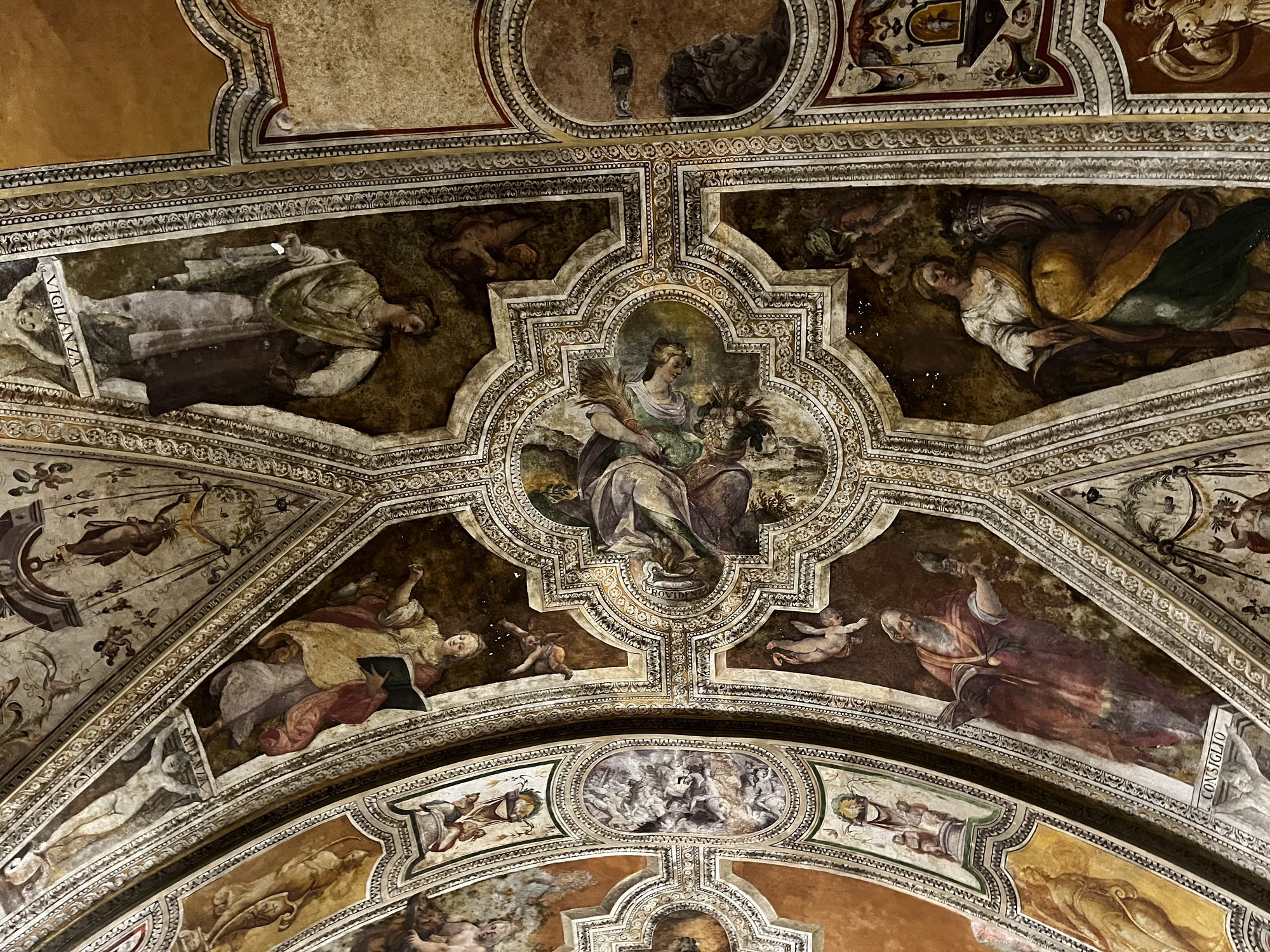 A close-up of an ornate frescoed vault inside a Naples museum — gilded plaster, painted figures and quatrefoil panels in faded colour