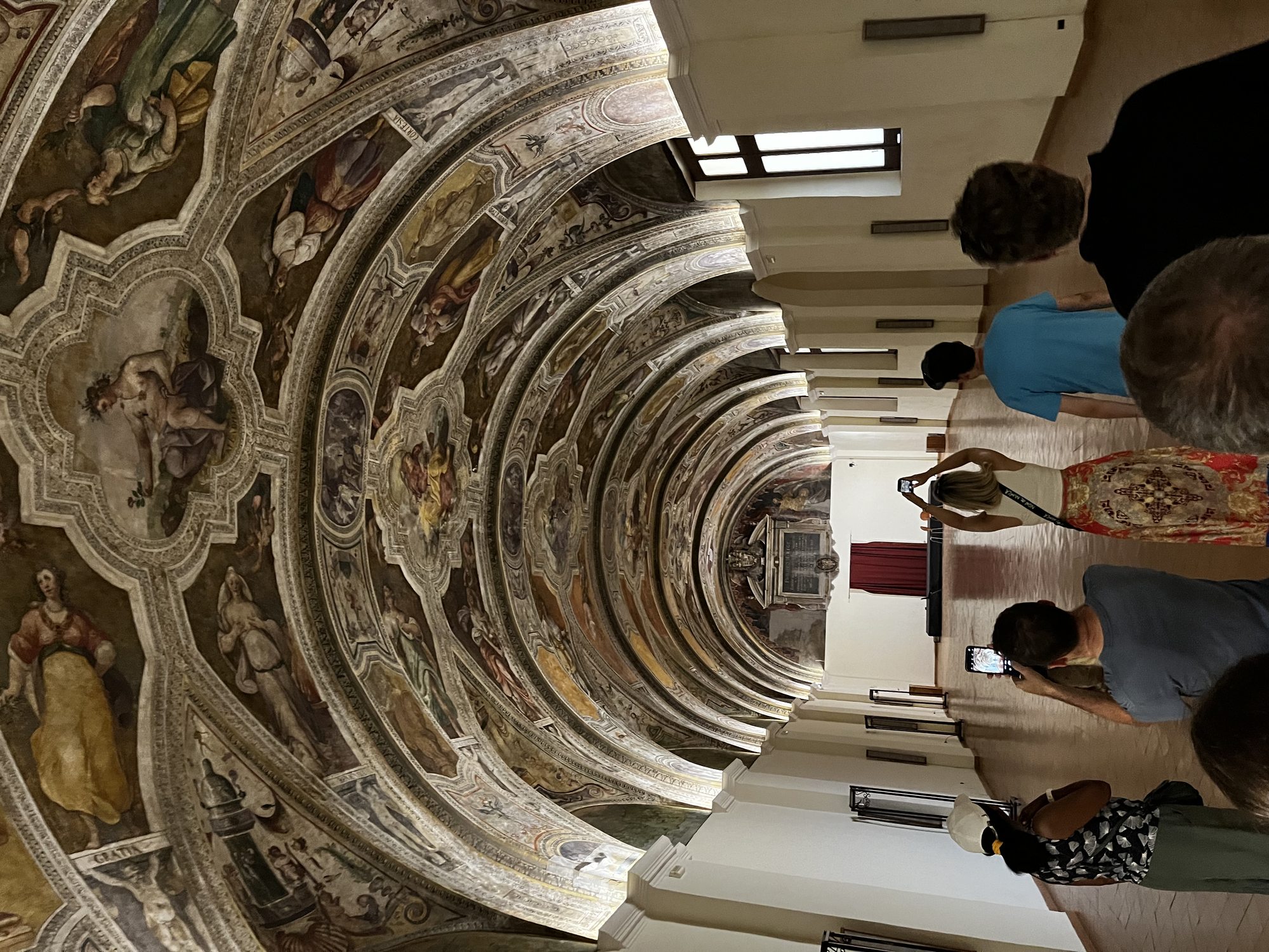 An ornate frescoed barrel vault inside a Naples museum — multiple painted panels stretching down a long corridor, visitors looking up in awe