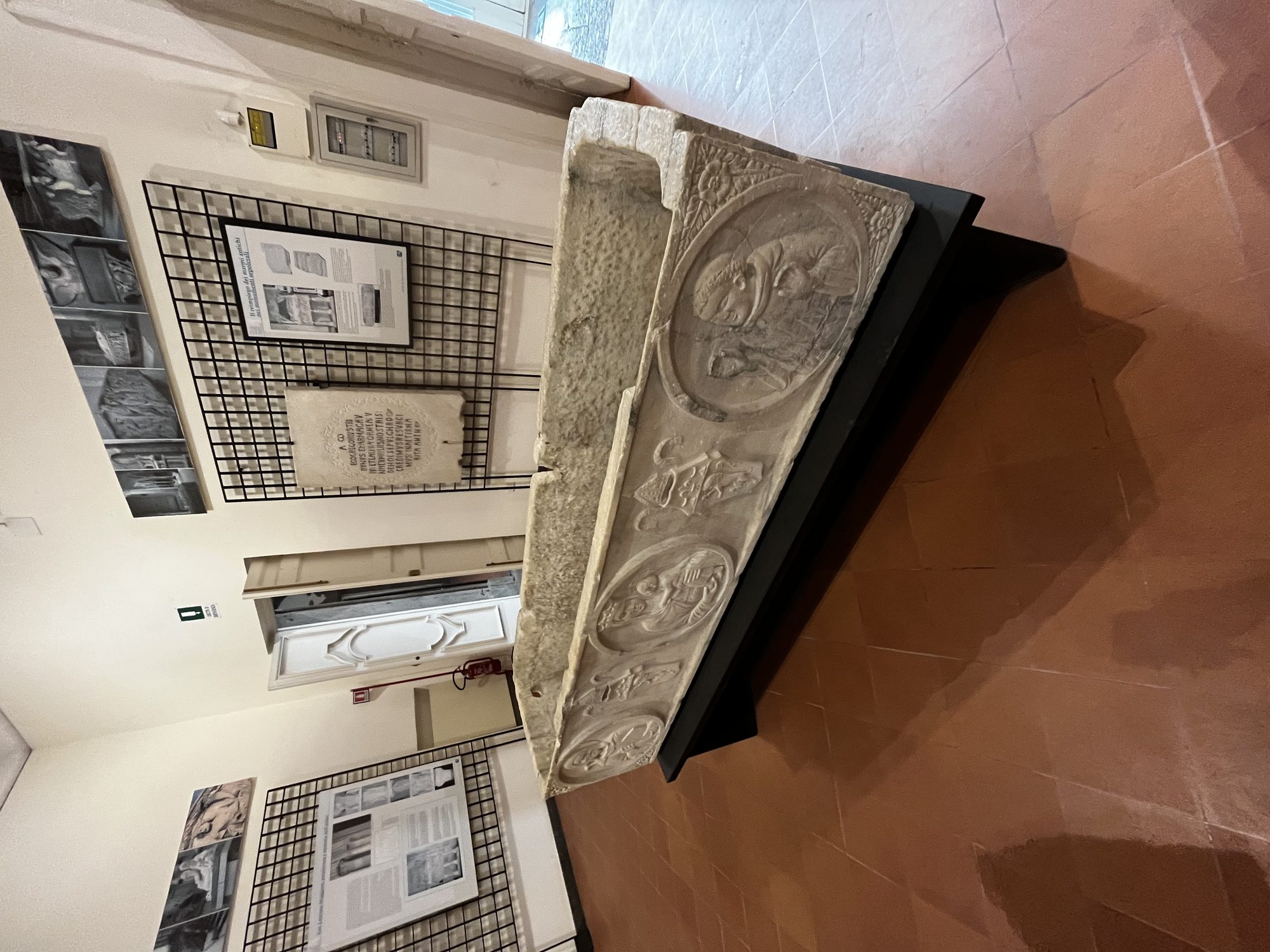 A Roman-era marble sarcophagus on display in the Naples Underground museum — carved reliefs, museum lighting, exhibition panels around it