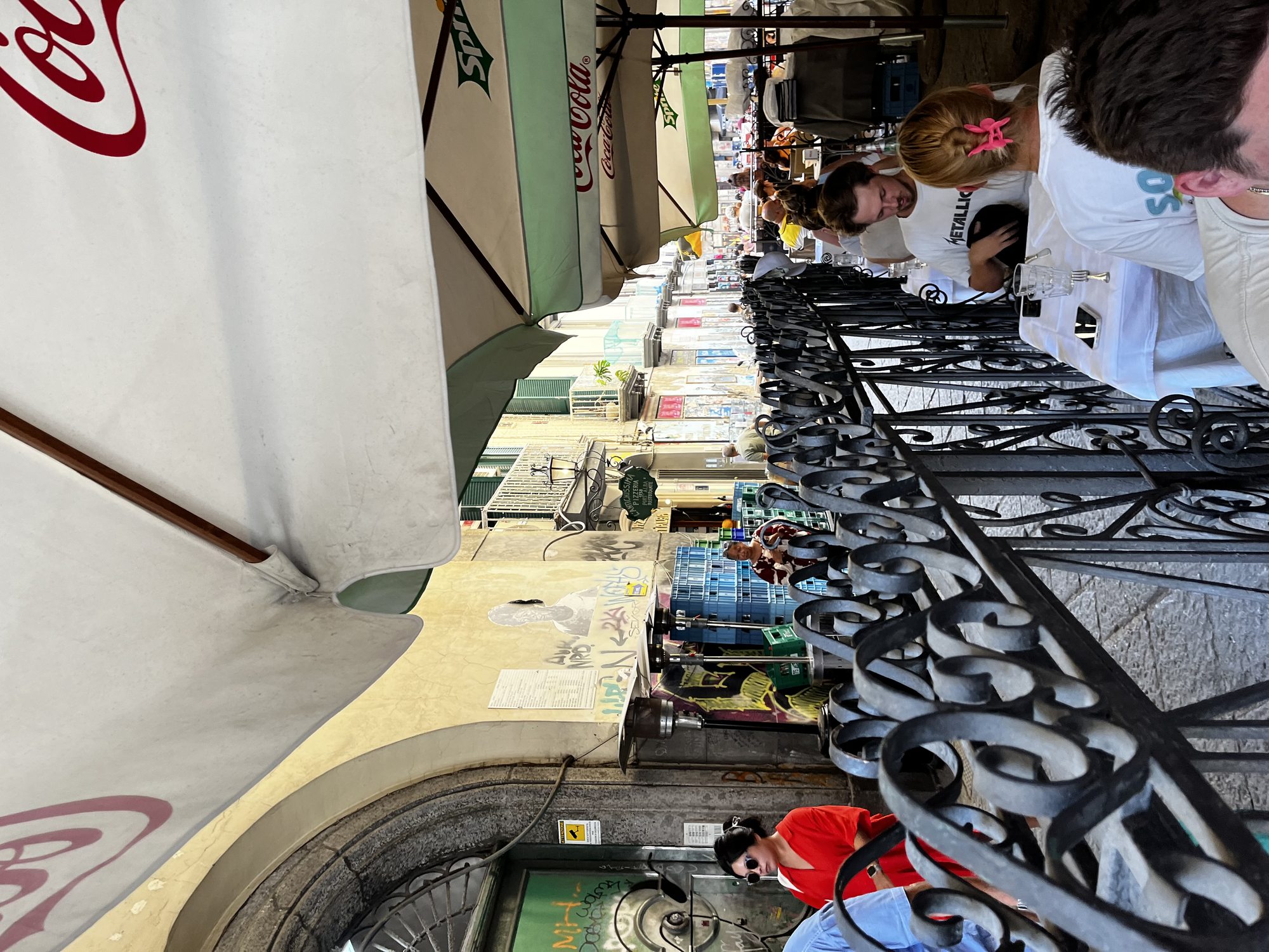 The covered terrace of Antica Pizzeria Port'Alba in Naples — long tables under Coca-Cola umbrellas, ironwork railings and the entrance of one of the world's oldest pizzerias visible at the far end