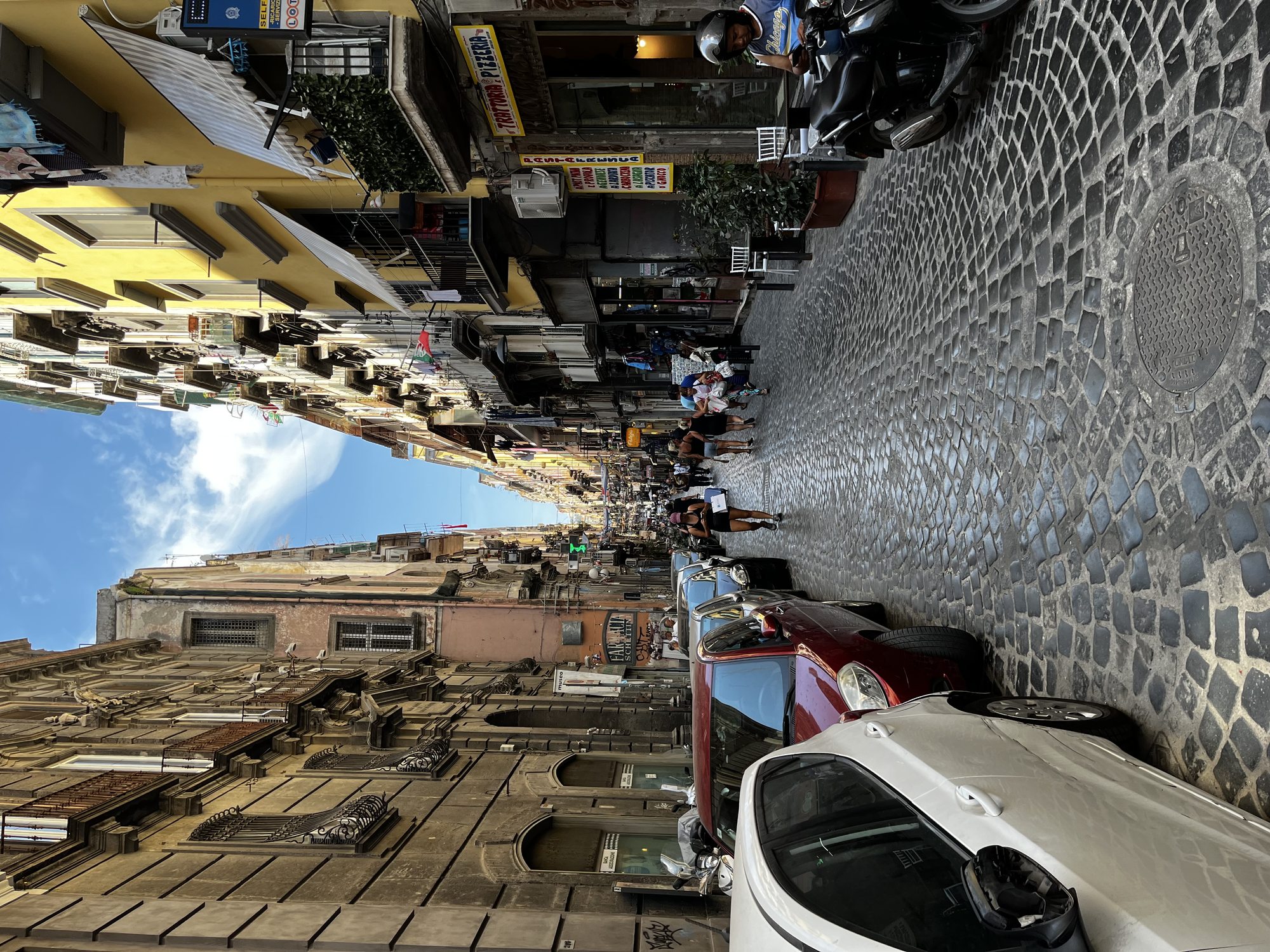 A narrow cobblestone street in the Quartieri Spagnoli — Naples' Spanish Quarters — with parked cars, market awnings and laundry above