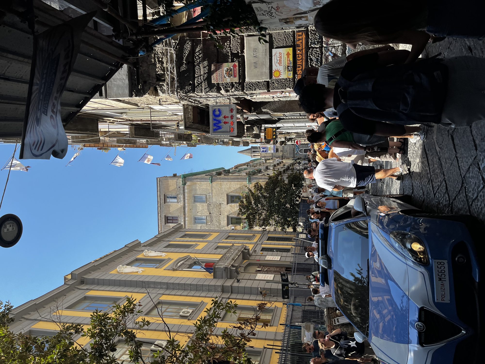 A busy street scene in central Naples — pedestrians, scooters, cars and a vivid mix of old and modern facades against an Italian summer sky