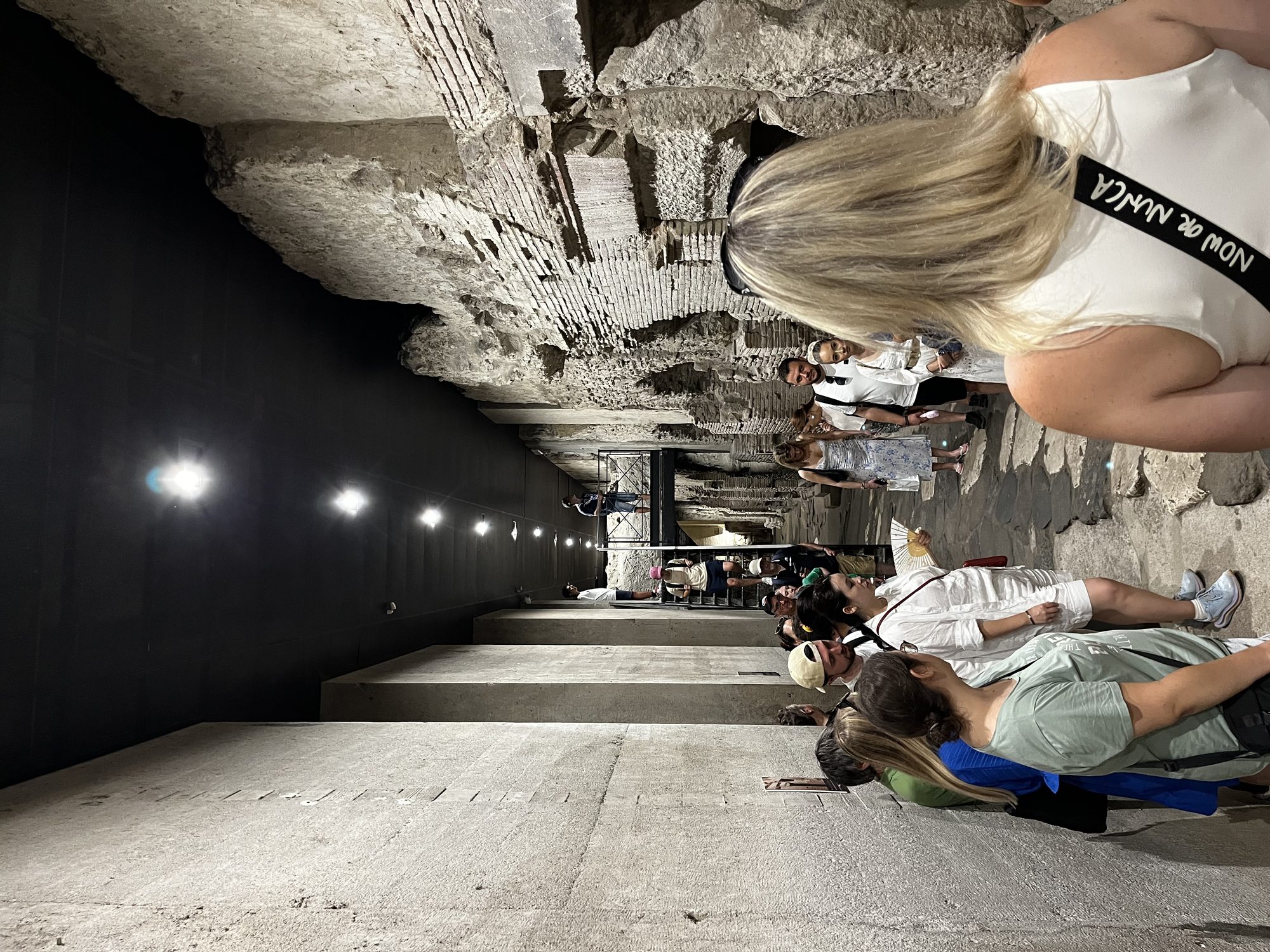 A vaulted underground chamber beneath Naples — visitors with phone torches gathered around the guide, ancient stone walls illuminated by warm light