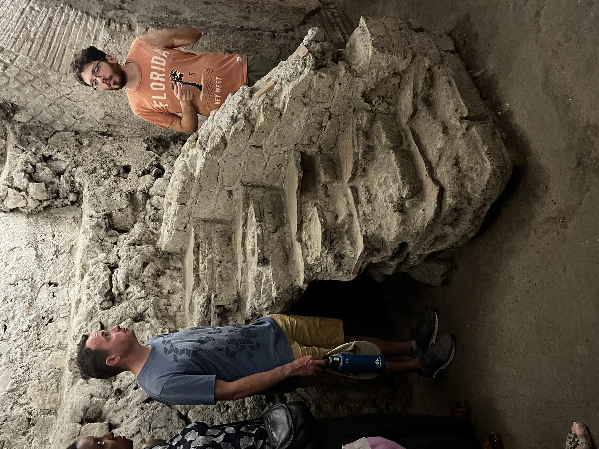 Two visitors examining a stone formation in a wide tuff-cut chamber deep beneath Naples — Greek-era quarry walls visible behind them