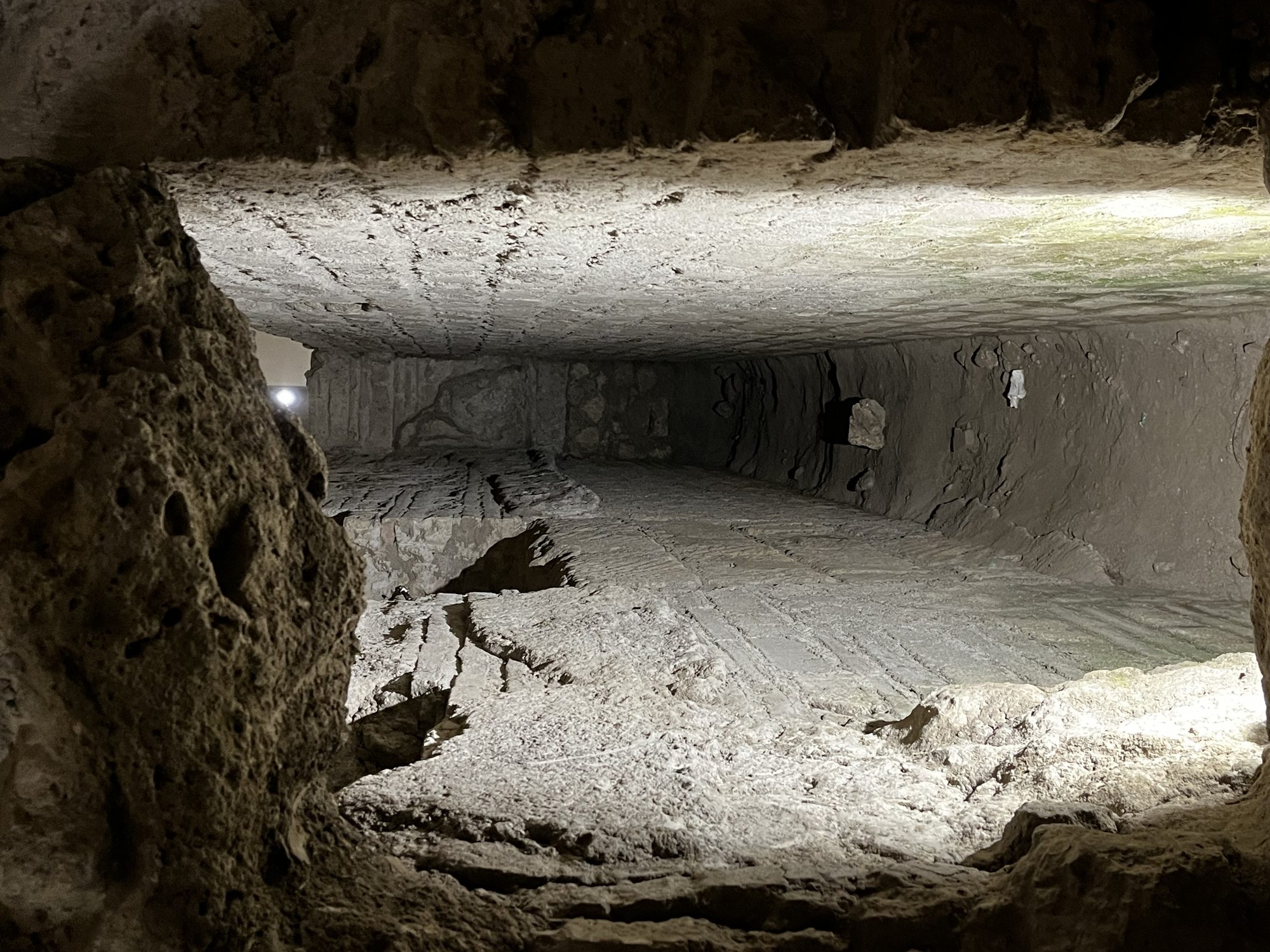 A long, raw stone tunnel deep beneath Naples — empty, cinematic, lit by a single warm light at the far end