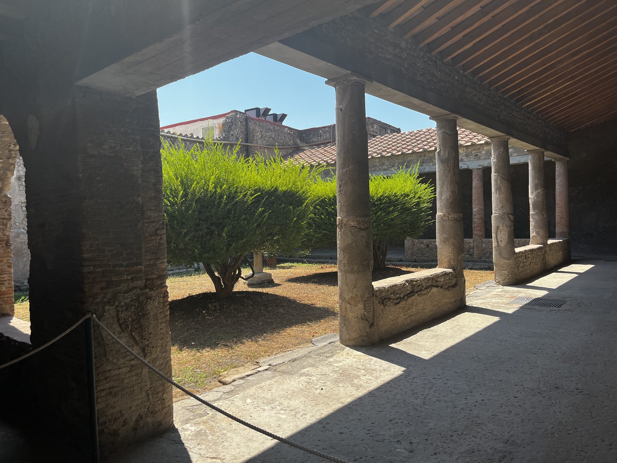 A shaded colonnade courtyard in Pompeii — stone columns framing a garden of green shrubs, the only shade in the ancient city