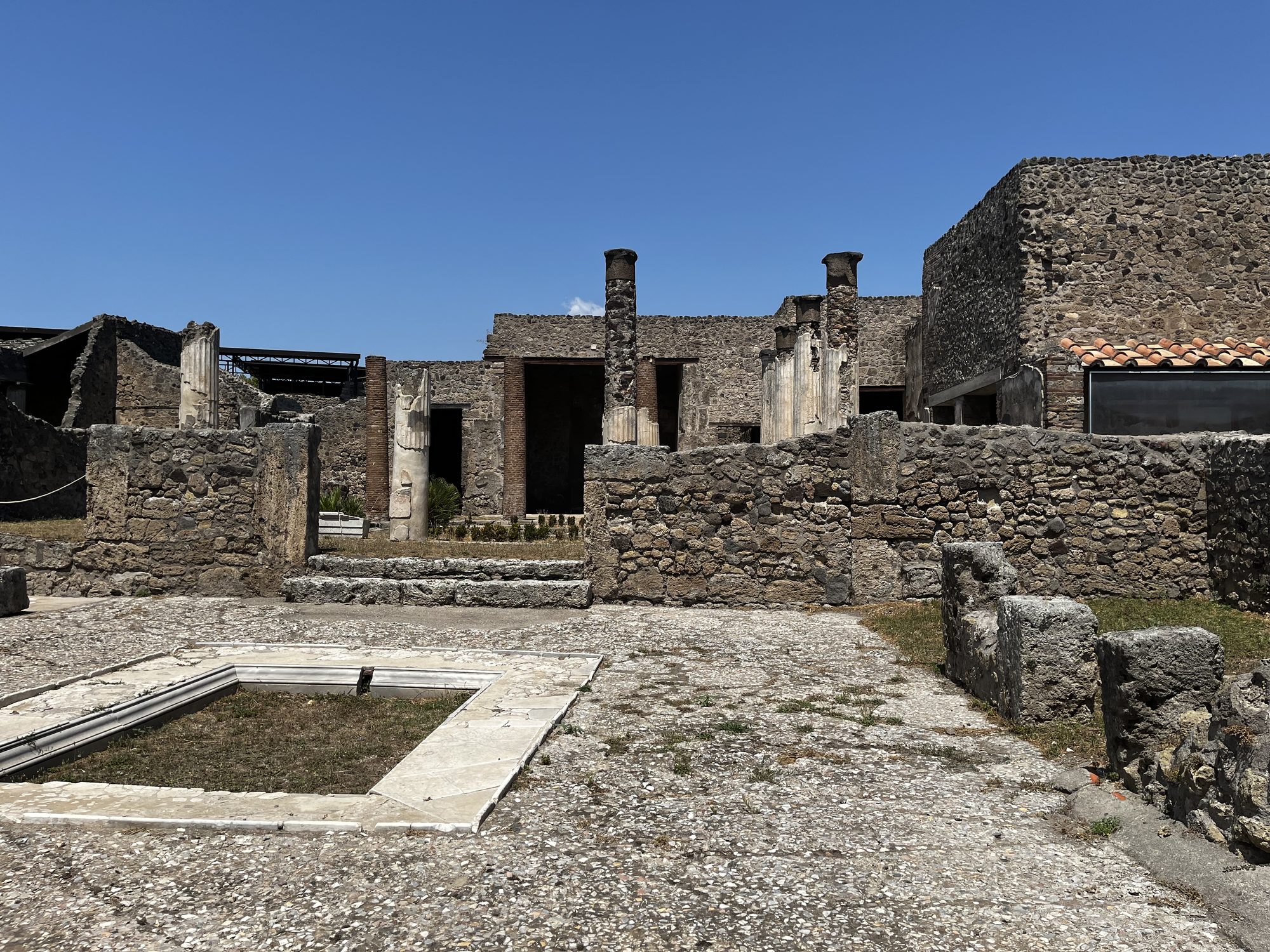 Open roofless ruins at Pompeii — the atrium of an ancient Roman house with an impluvium pool at the centre, Vesuvius visible beyond the walls