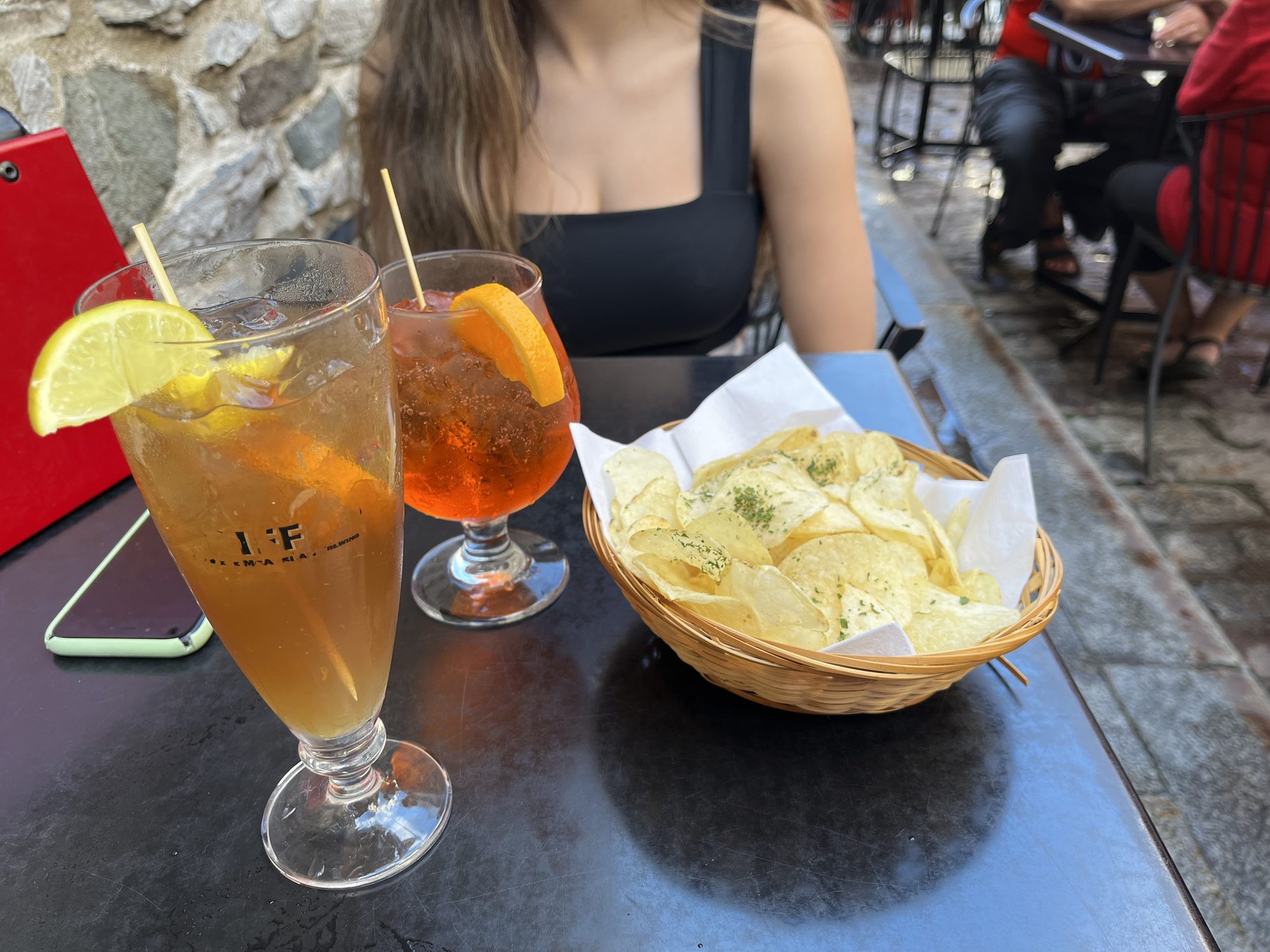An outdoor terrace table with a tall pilsner glass of pale beer with a lemon slice, an Aperol Spritz in a balloon glass with an orange slice, and a wicker basket of seasoned potato chips, with the back of a woman in a black dress sitting across the table