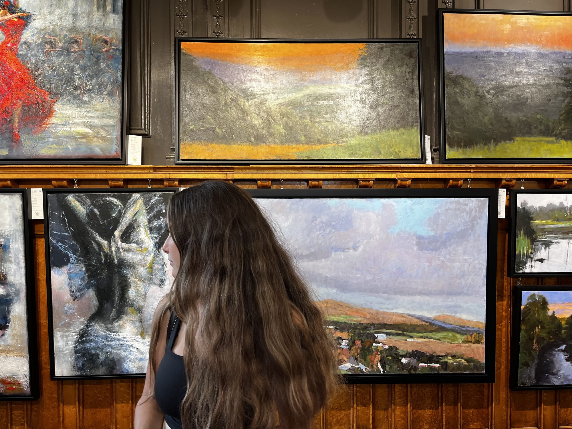 A young woman with long brown hair looking at oil paintings hung on a wood-panelled wall inside a Vieux-Québec art gallery — landscapes and figurative paintings stacked in rows