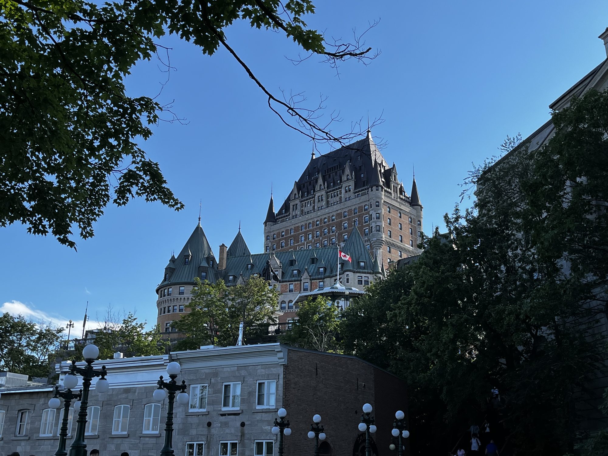 The Chateau Frontenac hotel from below — its green copper roof, multiple turrets and Canadian flag standing against a clear deep blue sky, framed by trees in the foreground
