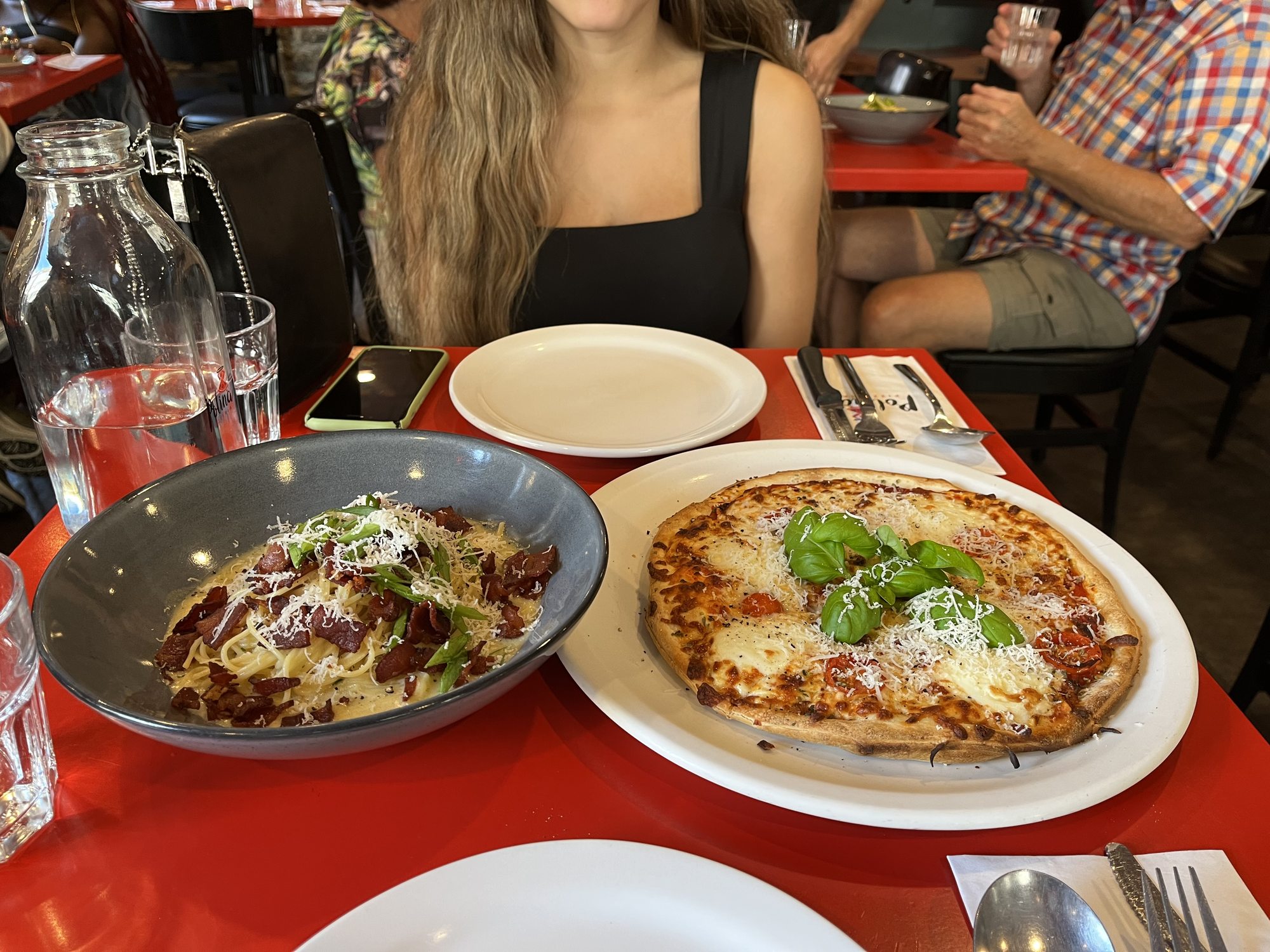 A dinner table with a deep blue bowl of pappardelle pasta with red meat sauce and parmesan, alongside a wood-fired pizza topped with melted mozzarella, basil leaves and prosciutto, on a red tablecloth at a Quebec City Italian restaurant