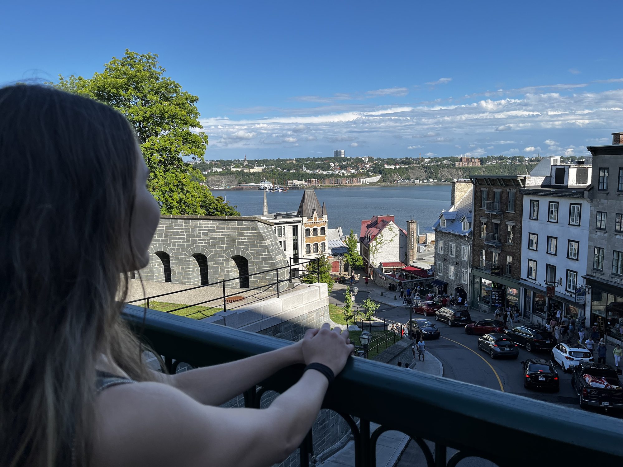 A view from Dufferin Terrace in Quebec City looking out over the Saint Lawrence river — the back of a woman with brown hair leaning on a green metal railing in the foreground, lower town buildings and the wide river beyond