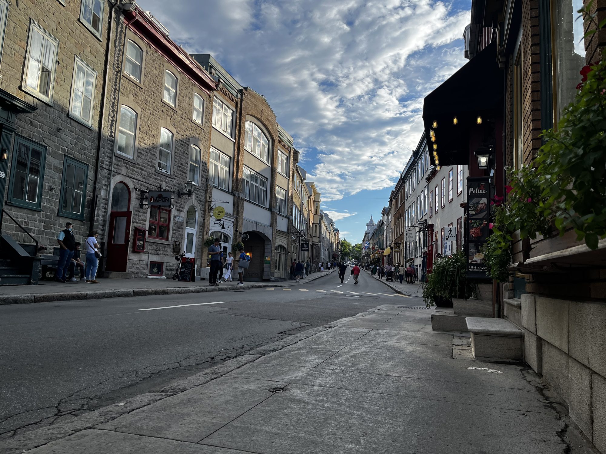 A long view down a sloping stone-paved street in Upper Town Quebec City in the early evening — stone buildings on either side, hanging shop signs, fewer pedestrians in the soft light, sky bright with light cloud cover