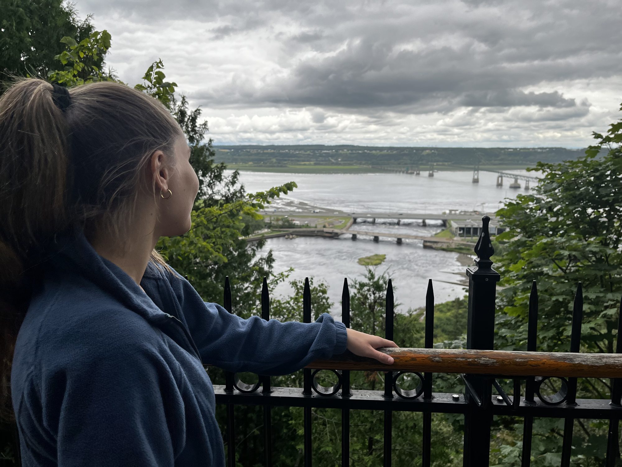 A woman in a blue sweatshirt leaning on an iron fence at the Chutes Montmorency lookout, looking out over the Saint Lawrence river with the Île d'Orléans bridge visible in the distance under grey cloud cover
