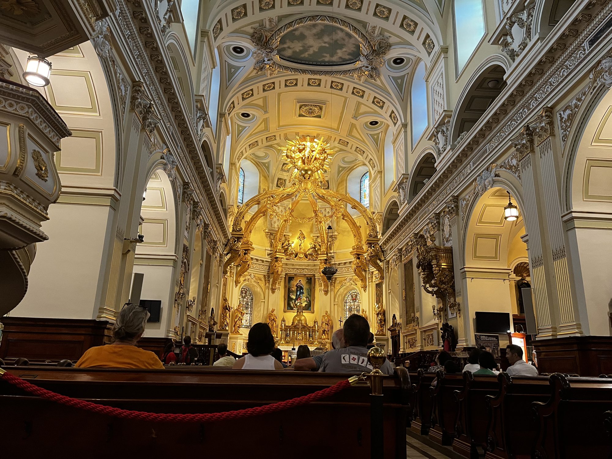 Interior of Basilique-Cathédrale Notre-Dame de Québec — view from the back of the nave toward the gilded baroque main altar, with the painted vaulted ceiling, ornate chandeliers, columns and worshipers seated in pews