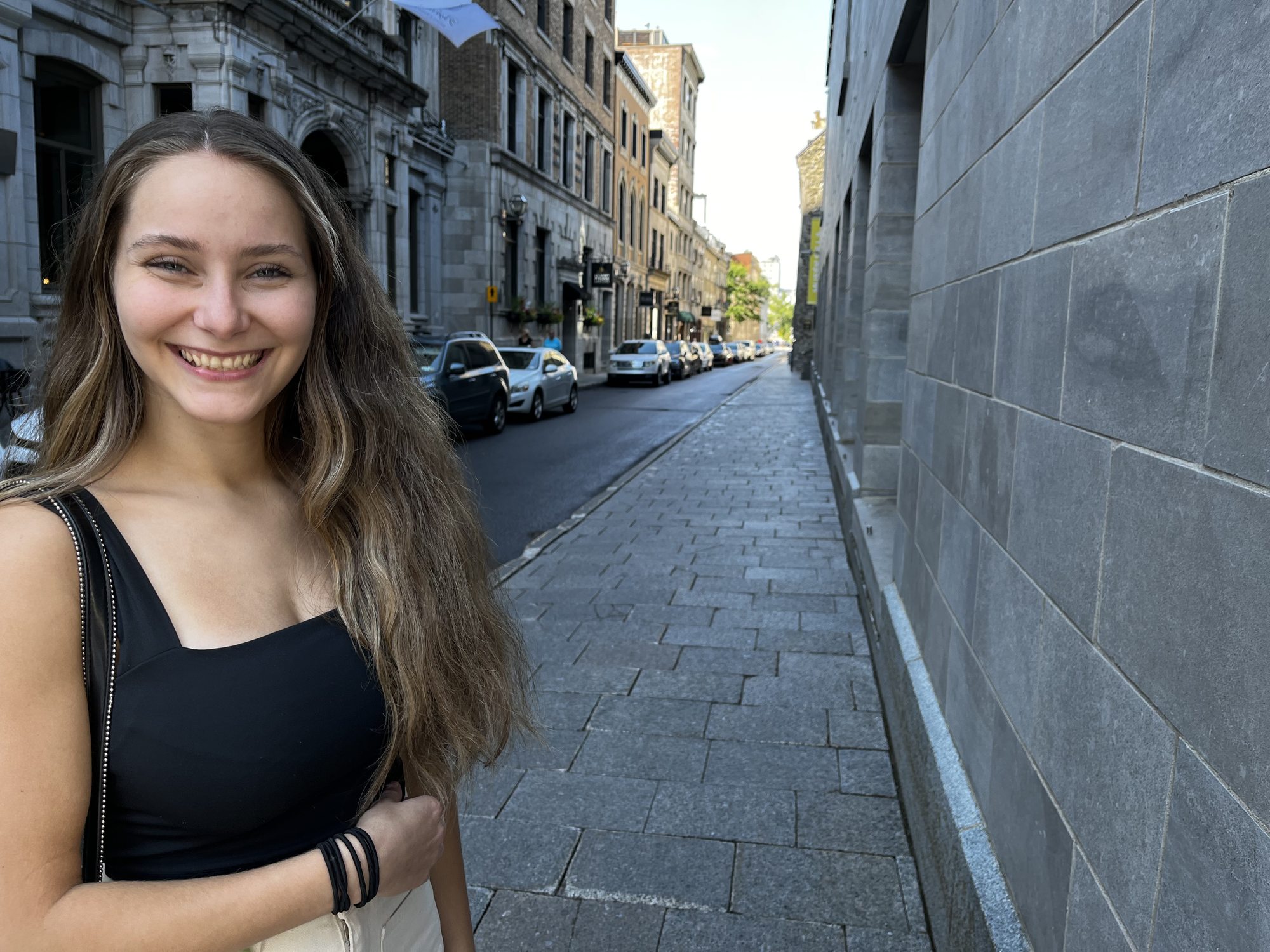 A young woman in a black top smiling on a stone-paved street in Vieux-Québec, with stone buildings, cars parked along a narrow street and an art gallery sign visible behind her