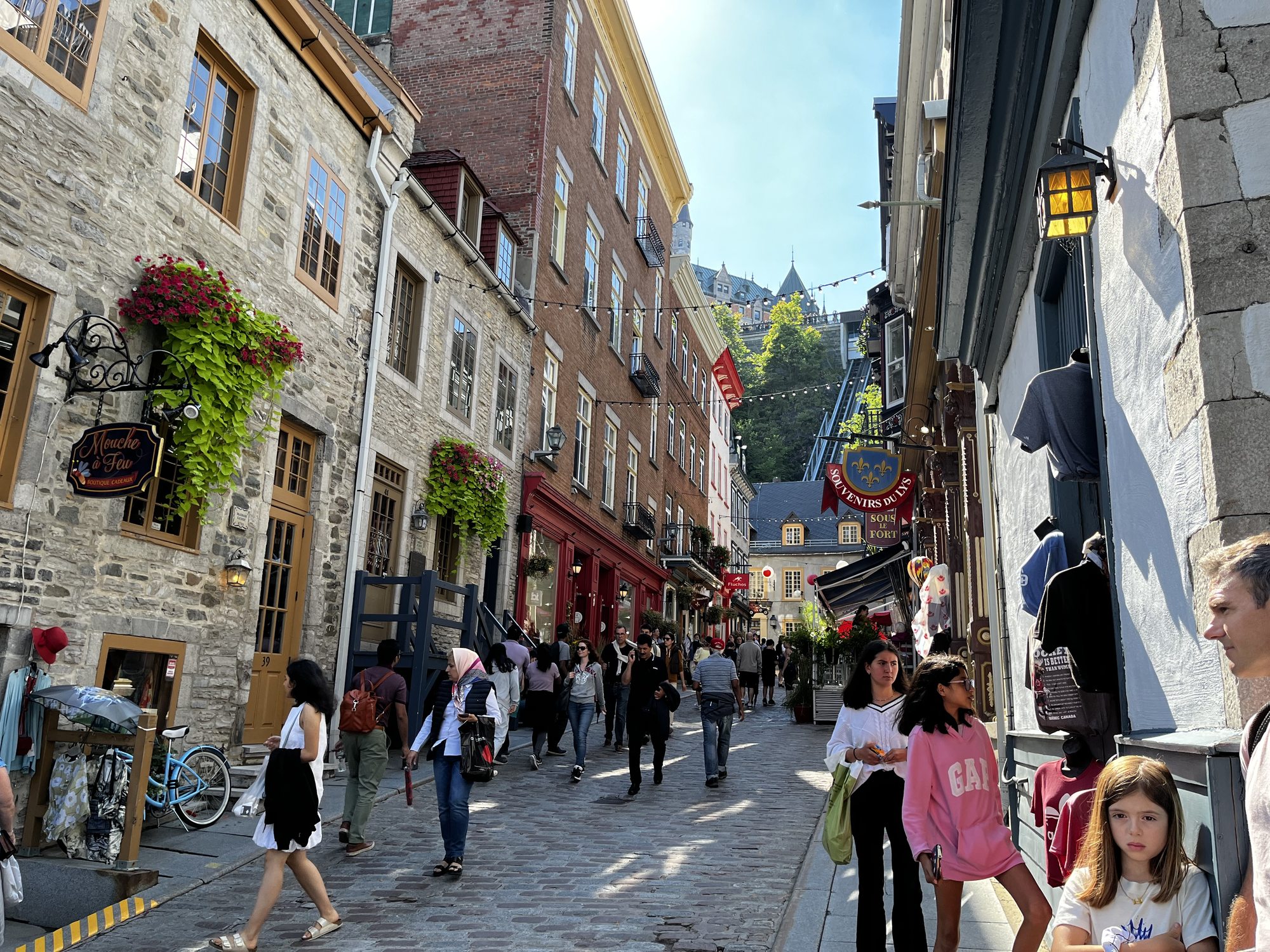 A crowded cobblestone street in Quartier Petit Champlain, Old Quebec, lined with stone buildings, hanging flower baskets and pub signs, with the cliff face and the funicular visible at the end of the street going up to the Chateau Frontenac