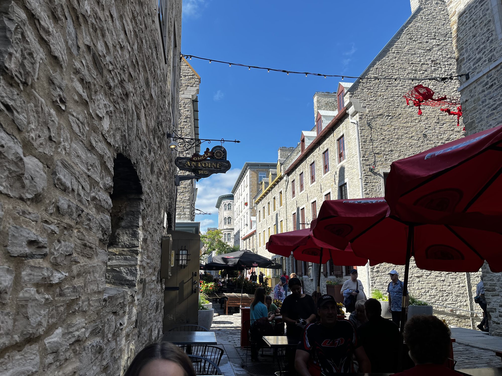 An alley in Old Quebec with stone walls on both sides, red café umbrellas set up over outdoor tables along the right side, an iron sign hanging from a building, blue sky overhead