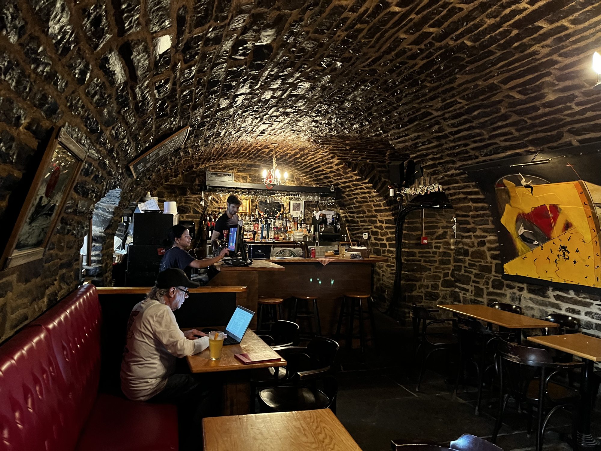 The vaulted stone interior of a historic Quebec City tavern — exposed brick arches overhead, a long wooden bar in the background, red leather banquettes, a stone wall with paintings hung on it, low warm lighting