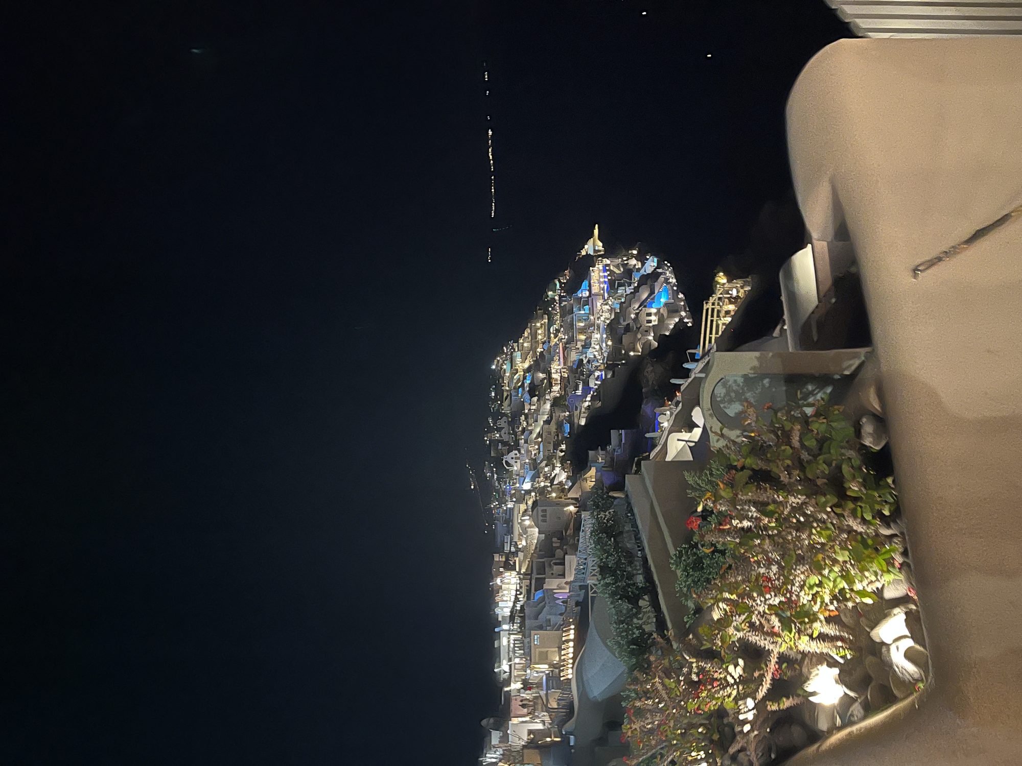 Santorini at night — the caldera villages lit up below, a woman sitting on the clifftop wall looking out
